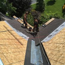 Deux couvreurs travaillent sur un toit, posant des bardeaux. Arbres verts et pelouse en arrière-plan.