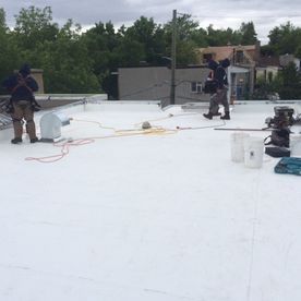 Deux ouvriers travaillent sur un toit plat blanc, probablement en train de le réparer. Des outils et des seaux sont visibles.