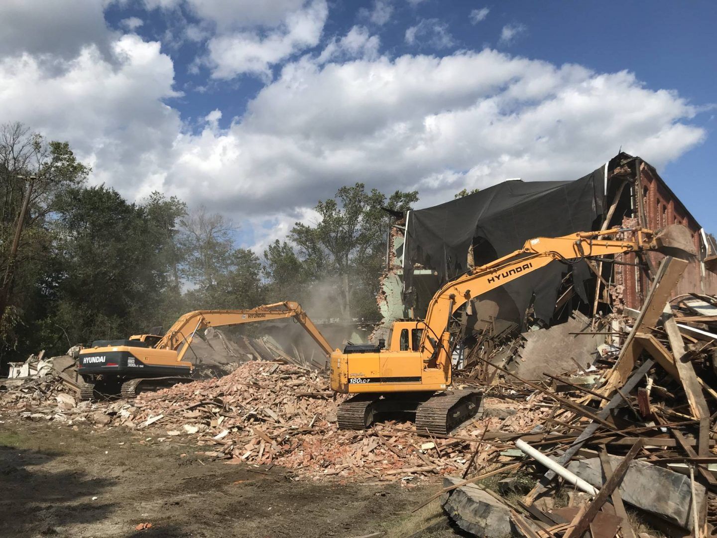 Two yellow excavators demolishing a brick building under a cloudy blue sky.