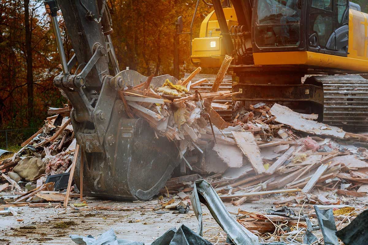 Excavator demolishing a structure, bucket filled with debris; yellow machine, outdoor setting.