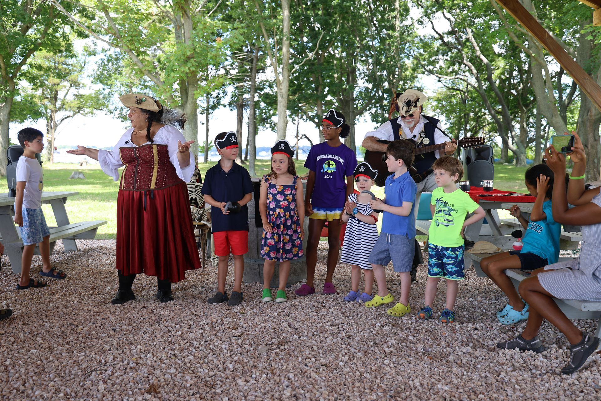 A costumed pirate leads a group of children at a park, while a musician plays in the background.