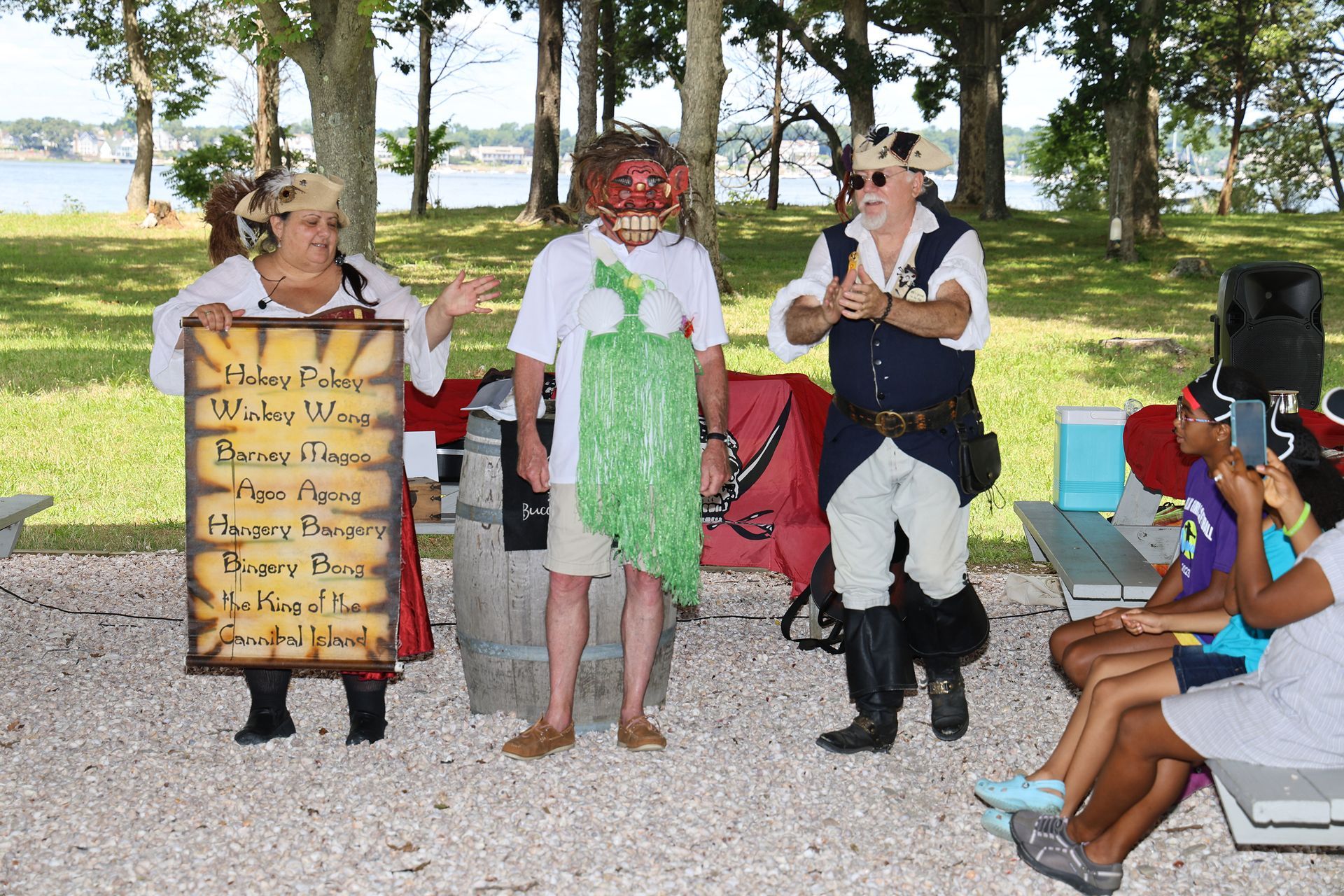 Three costumed people at an outdoor pirate-themed event, one holding a sign, others gesturing.