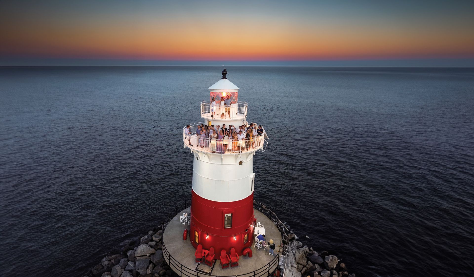 Lighthouse with red and white stripes, at dusk, with people on top, surrounded by dark water.