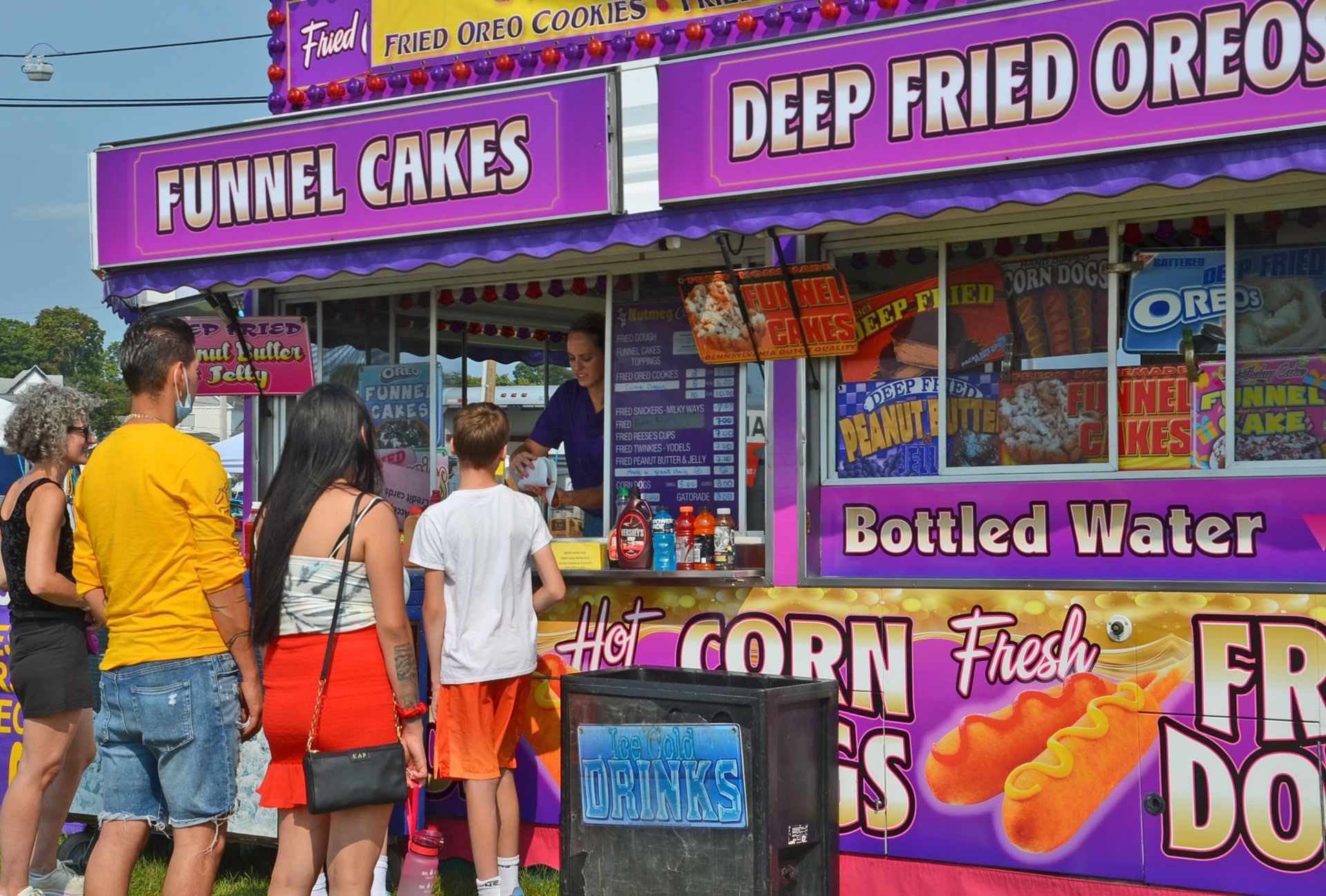Food stand with people in line; offers funnel cakes, deep-fried Oreos, corn dogs.