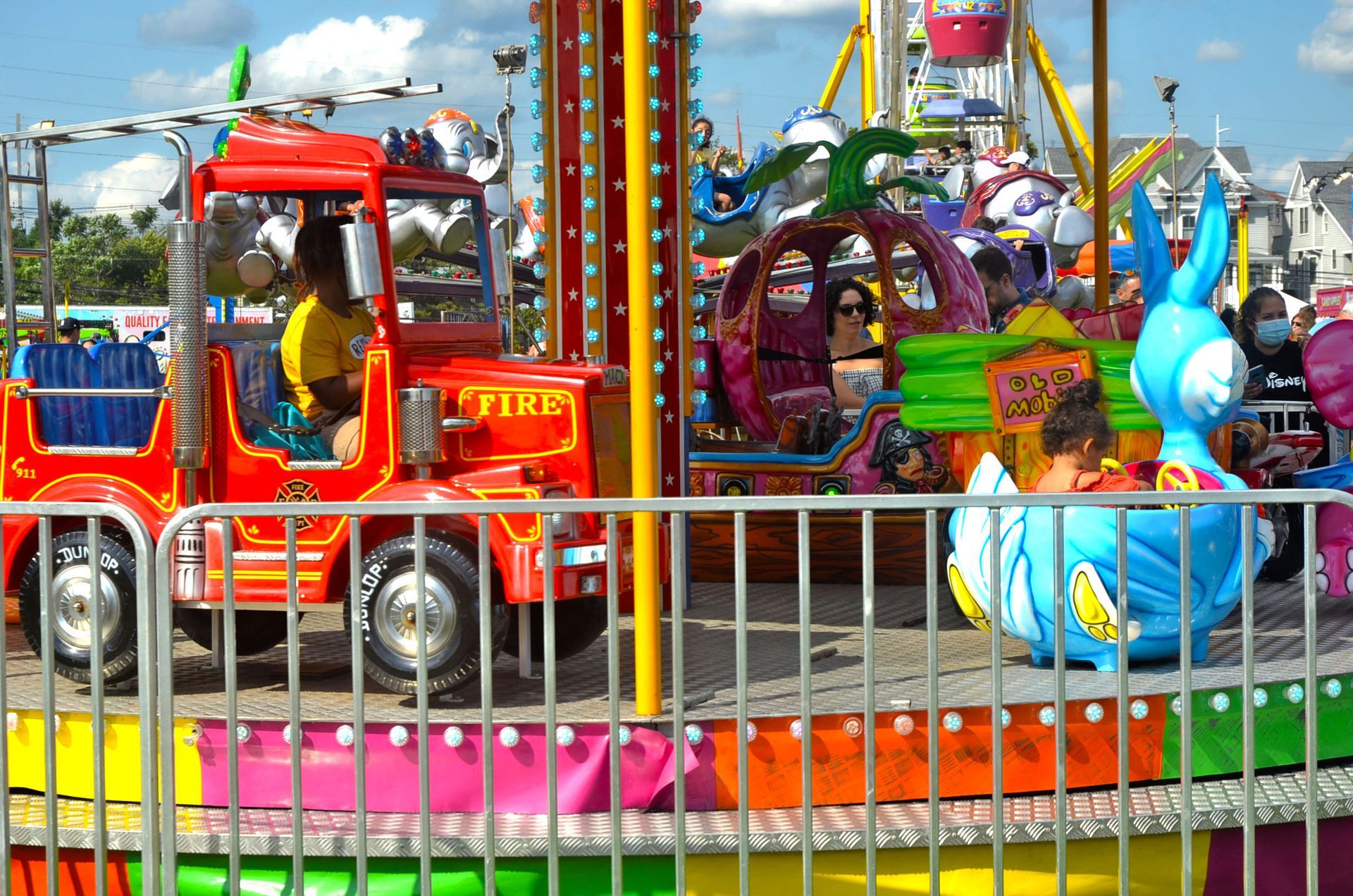 Carnival ride with fire truck, pumpkin, and duck-shaped cars on a carousel. Fence and Ferris wheel in background.