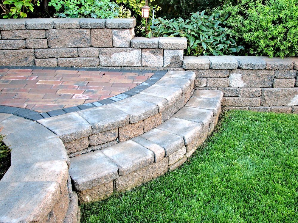 Stone retaining wall with curved steps leading to a brick patio. Green grass in the foreground.