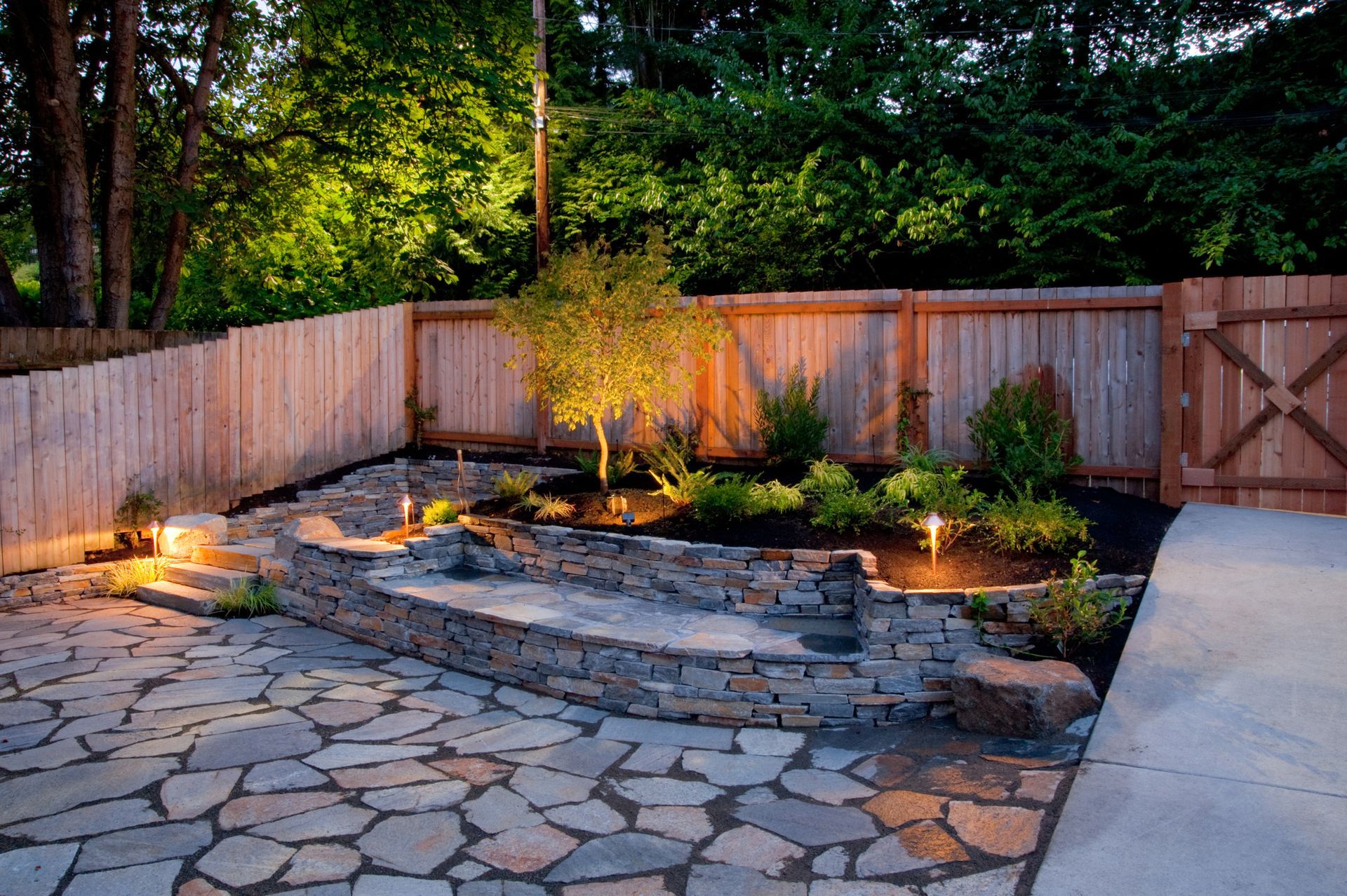 Backyard stone patio with a small raised pond, potted plants, and wooden fence at dusk