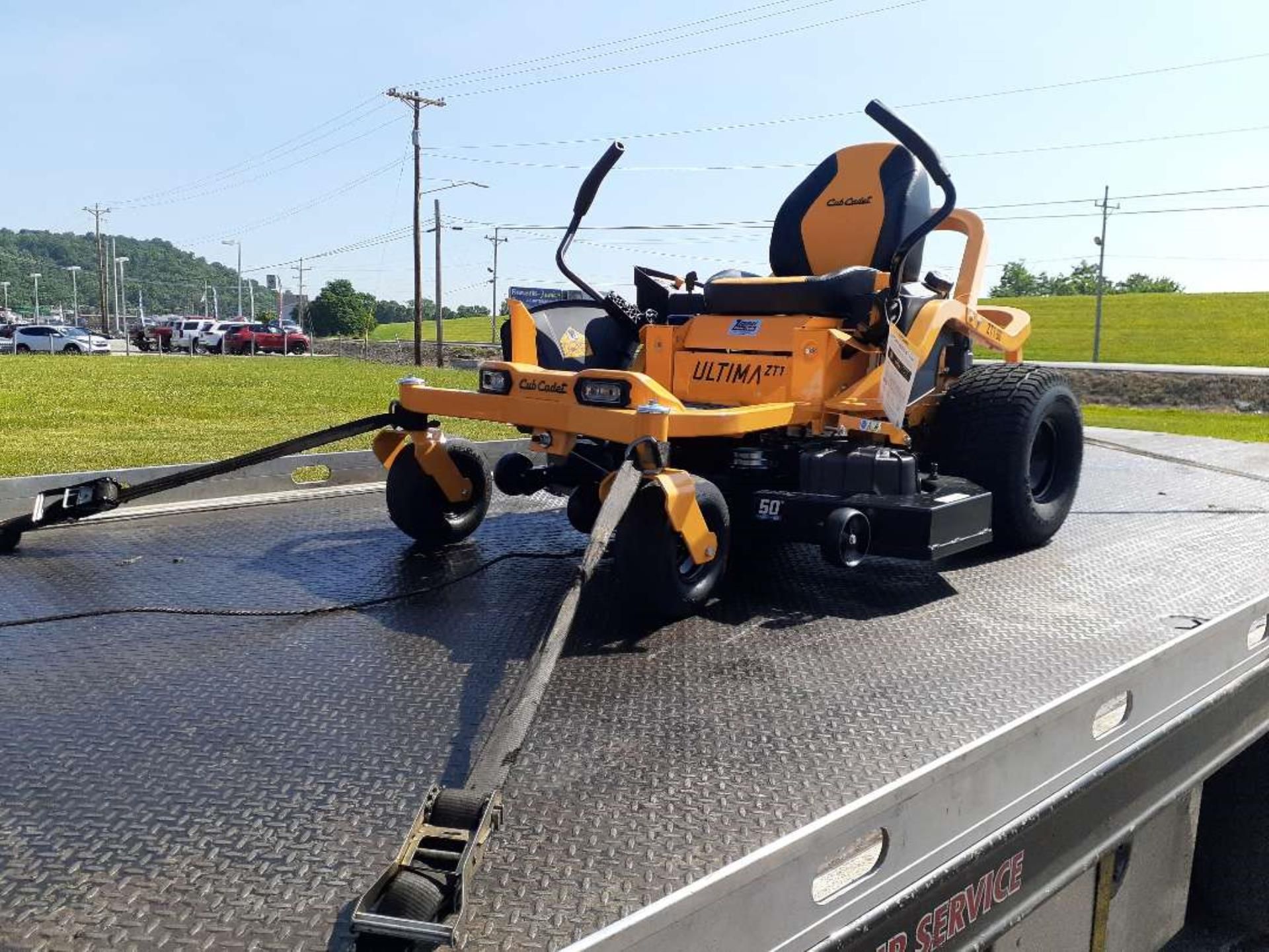 A yellow and black zero-turn lawn mower secured with straps on a flatbed trailer outdoors on a sunny day.
