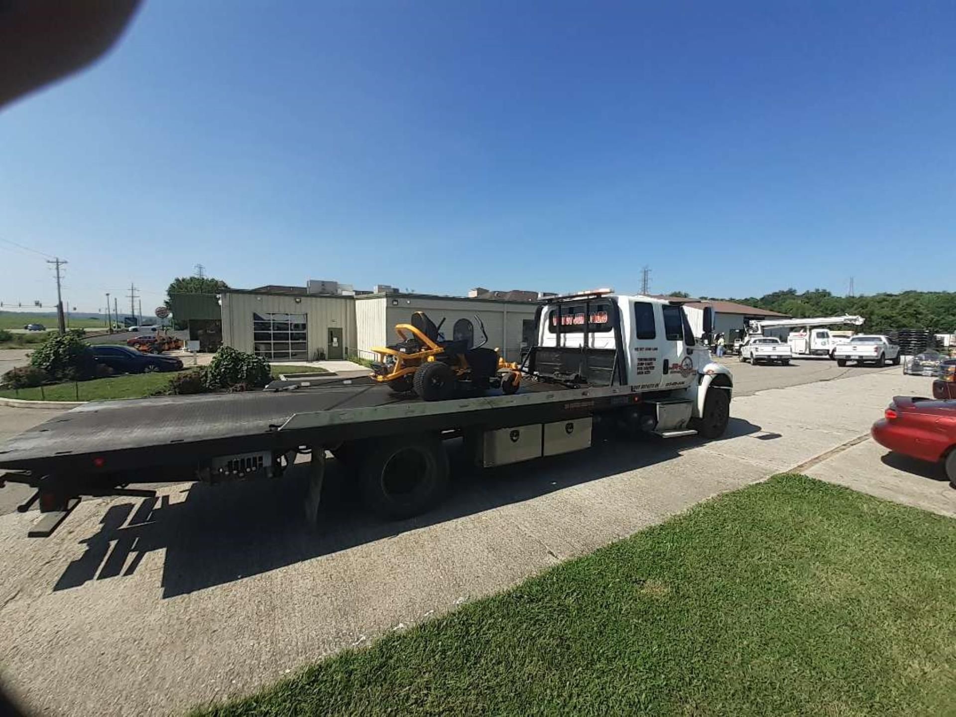 A white flatbed tow truck is parked on a gravel lot with a yellow lawn mower secured on its bed under a blue sky.