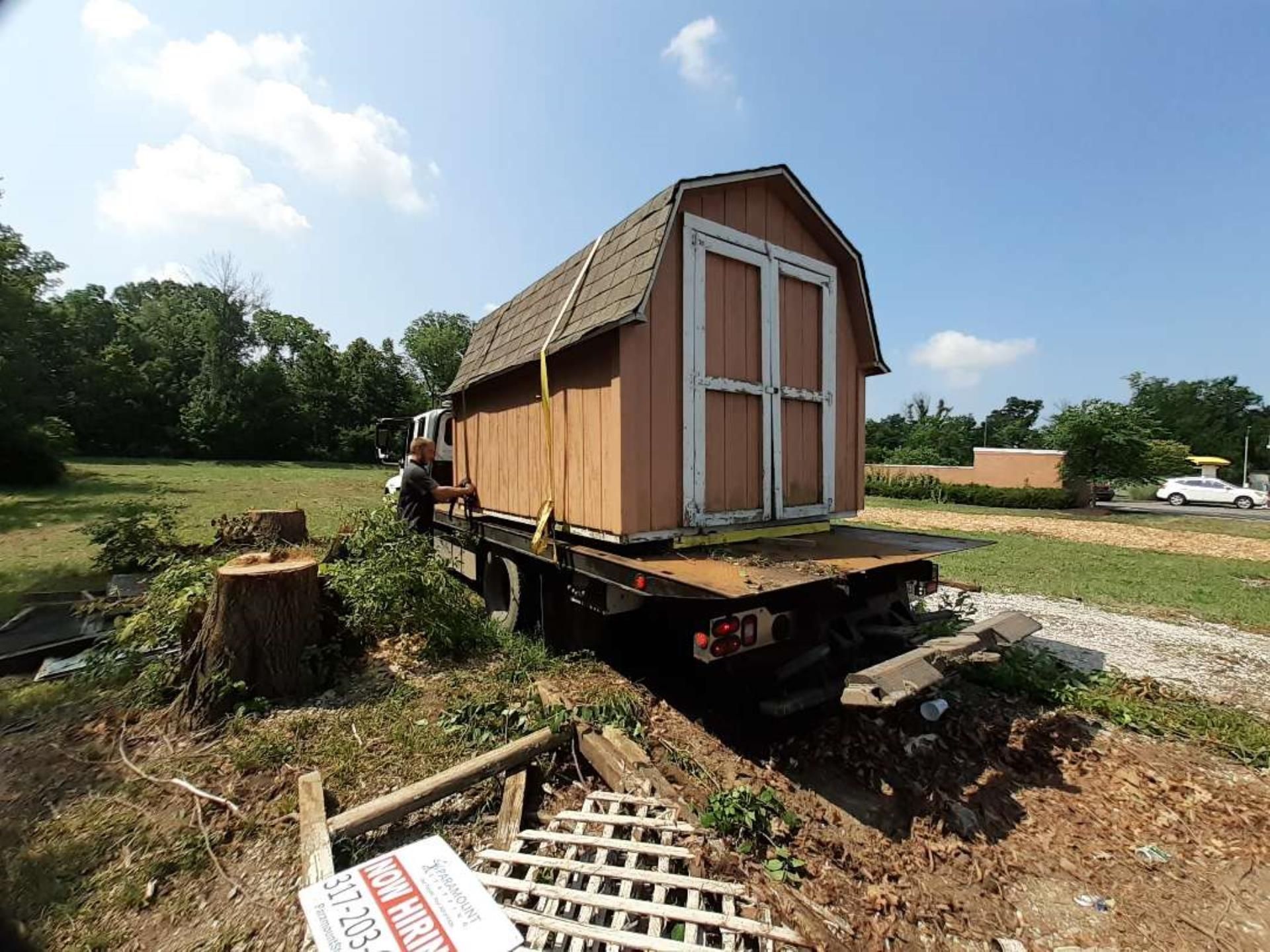 A tan storage shed sits on the flatbed of a parked truck in a grassy field, secured with yellow nylon straps.