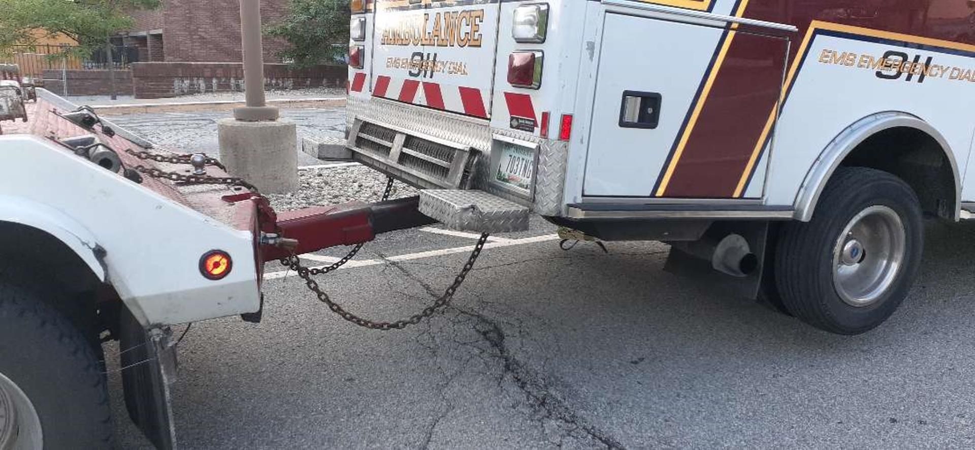 A white tow truck is towing an ambulance on a paved road.