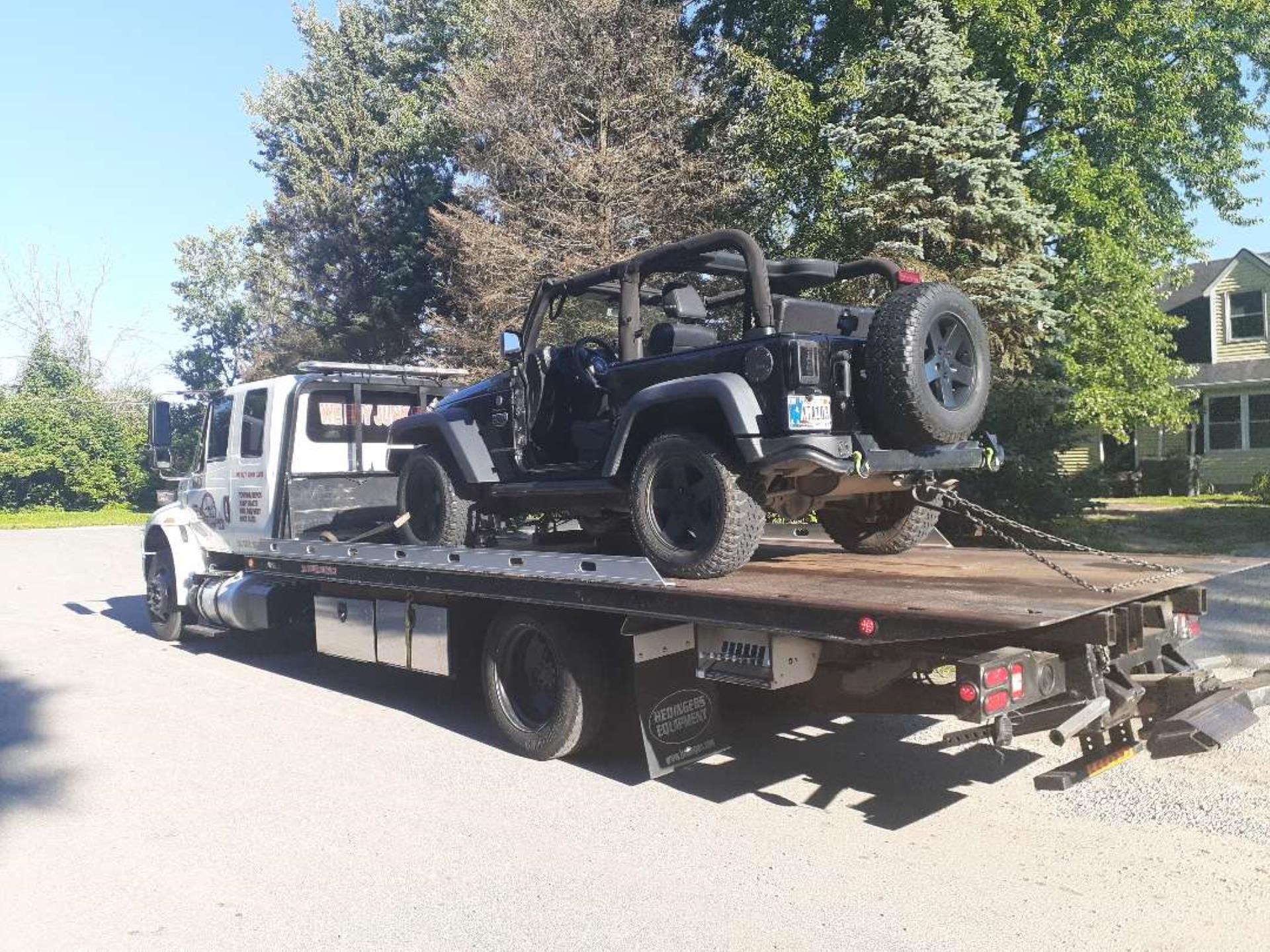 A black Jeep being transported on the flatbed of a white tow truck parked on a gravel road.