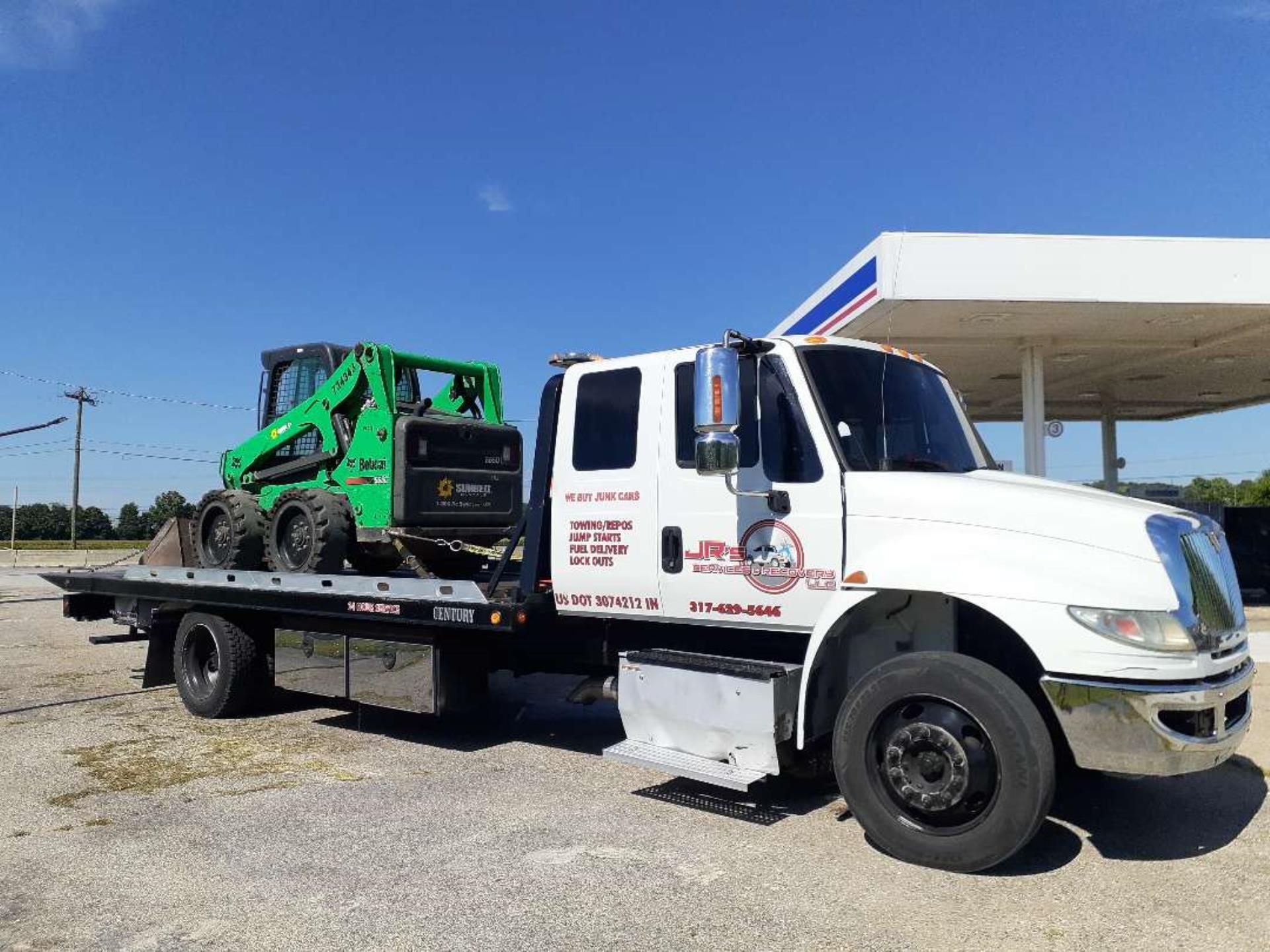 A white flatbed tow truck carrying a green skid-steer loader parked near a gas station.