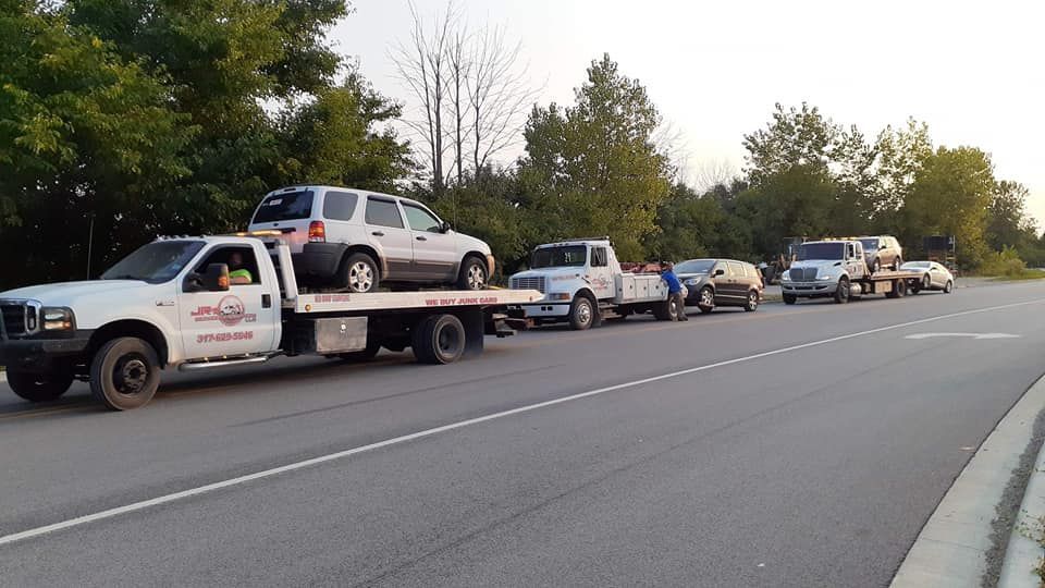 A line of flatbed tow trucks parked on a road, each carrying a vehicle, with green trees in the background.