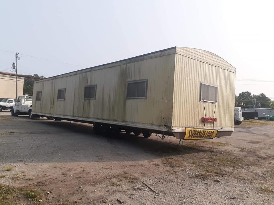 A long, beige mobile office trailer sits on a gravel lot under a bright, overcast sky.