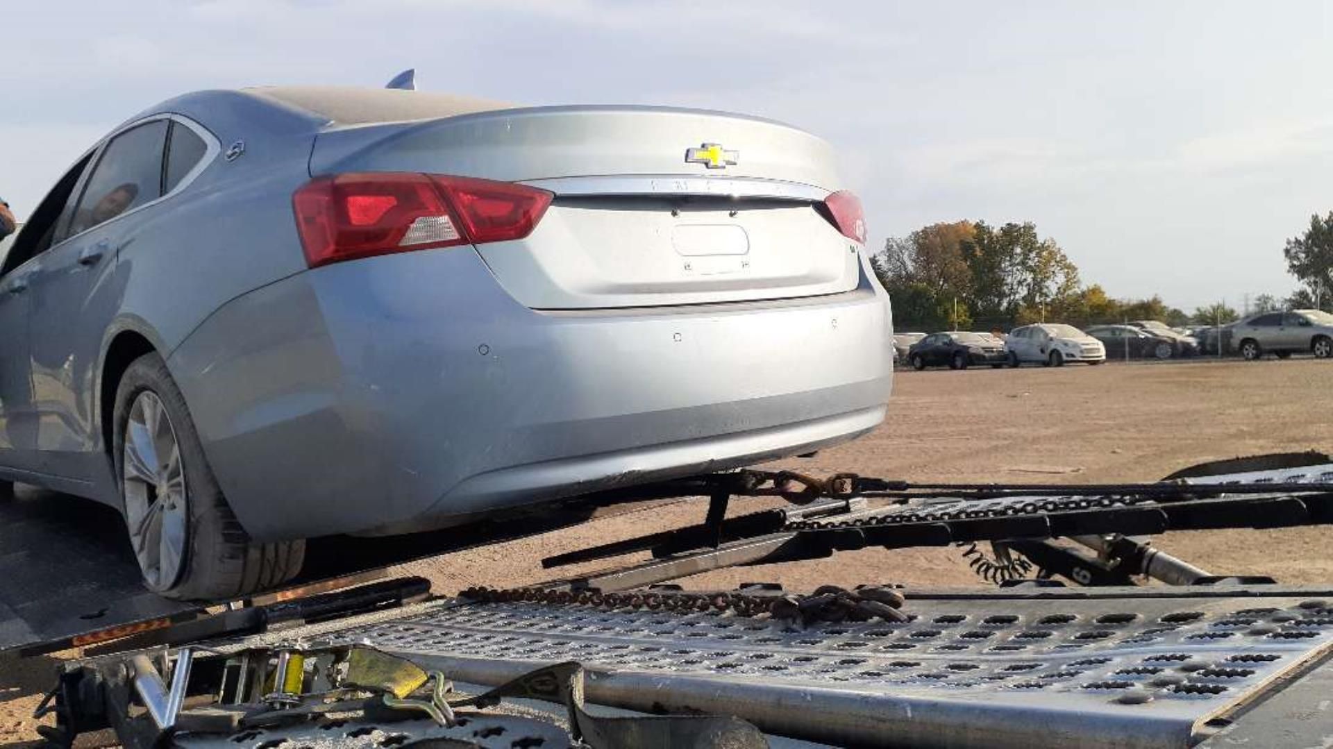 A light blue Chevrolet sedan is being transported on a flatbed trailer in a dirt lot.