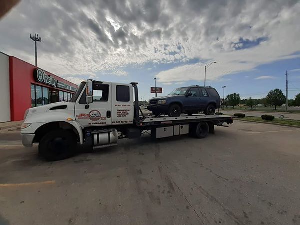 A white tow truck parked in a lot carrying a dark blue SUV on its flatbed under a cloudy sky.
