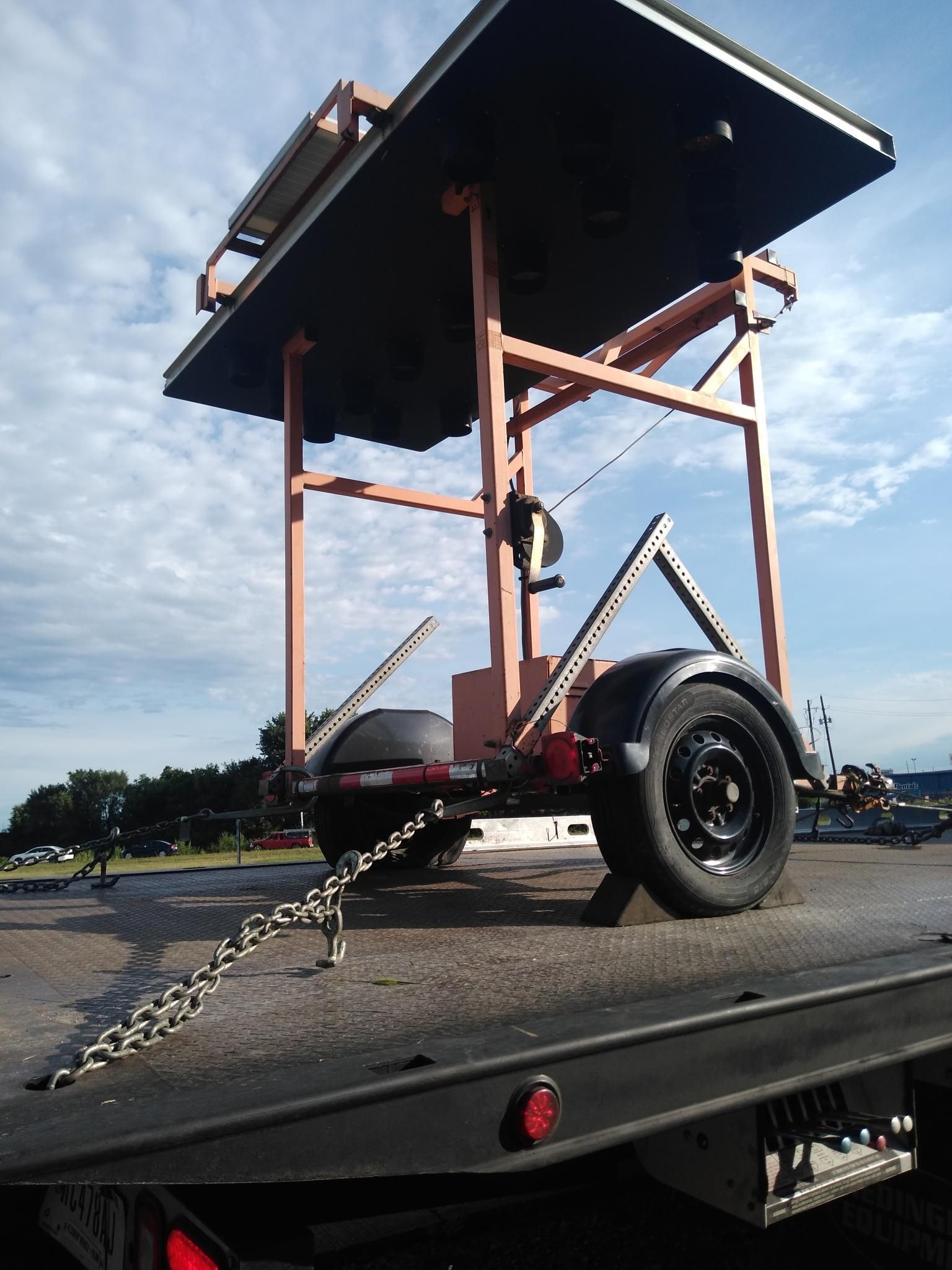 A light-orange metal frame structure is secured by chains to the bed of a flatbed truck outdoors under a blue sky.