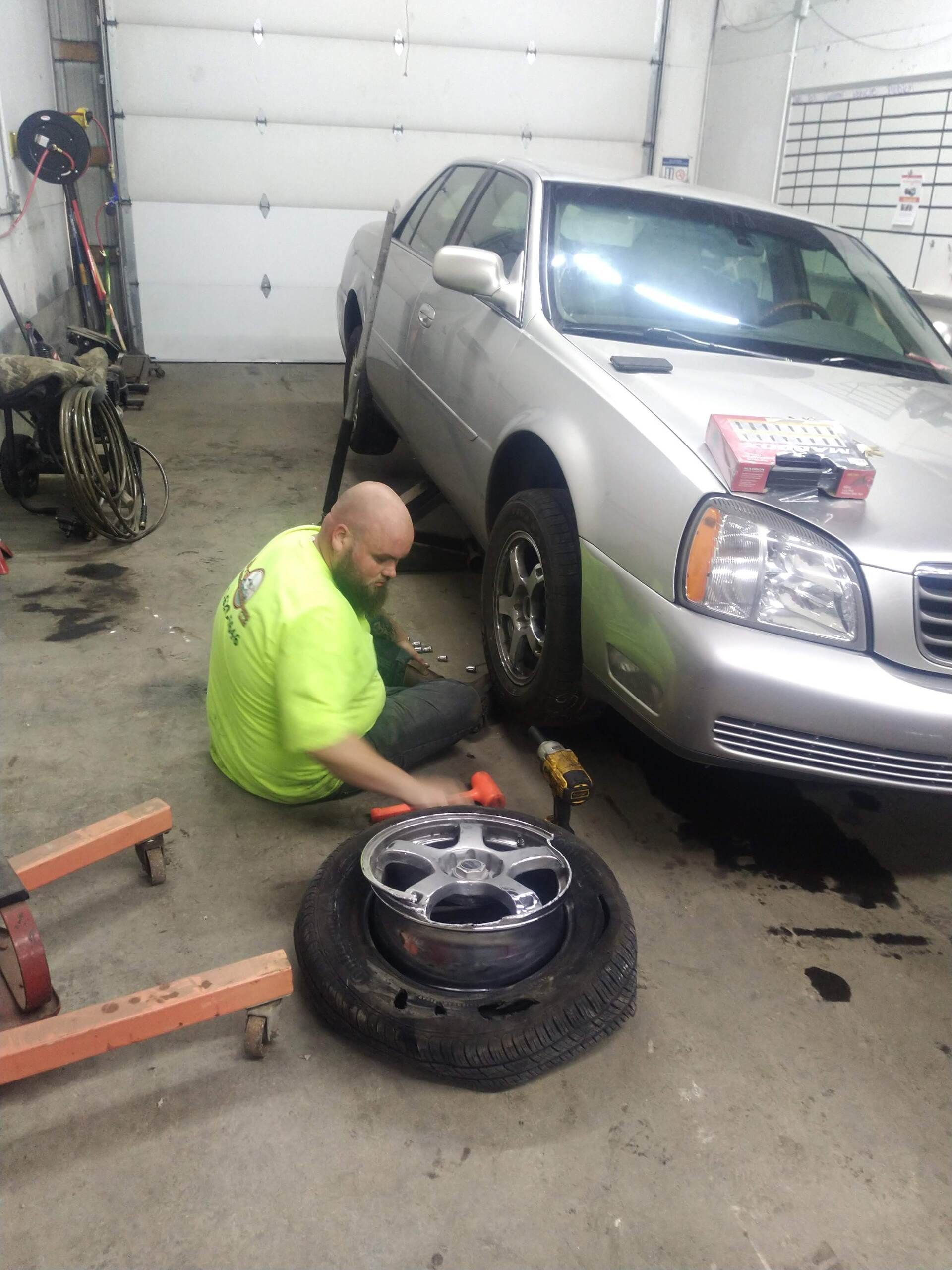 A mechanic in a bright yellow shirt uses a hammer to work on a car lifted on a jack in an auto repair shop.