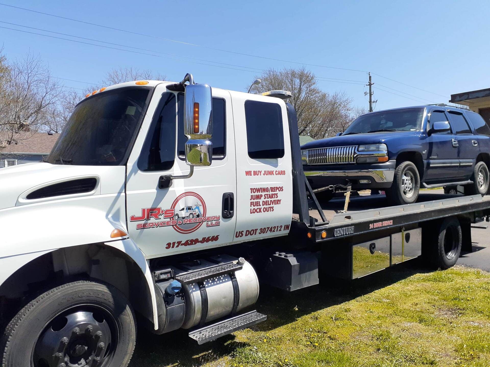 A white J&R towing truck carrying a dark blue SUV on its flatbed against a clear blue sky.