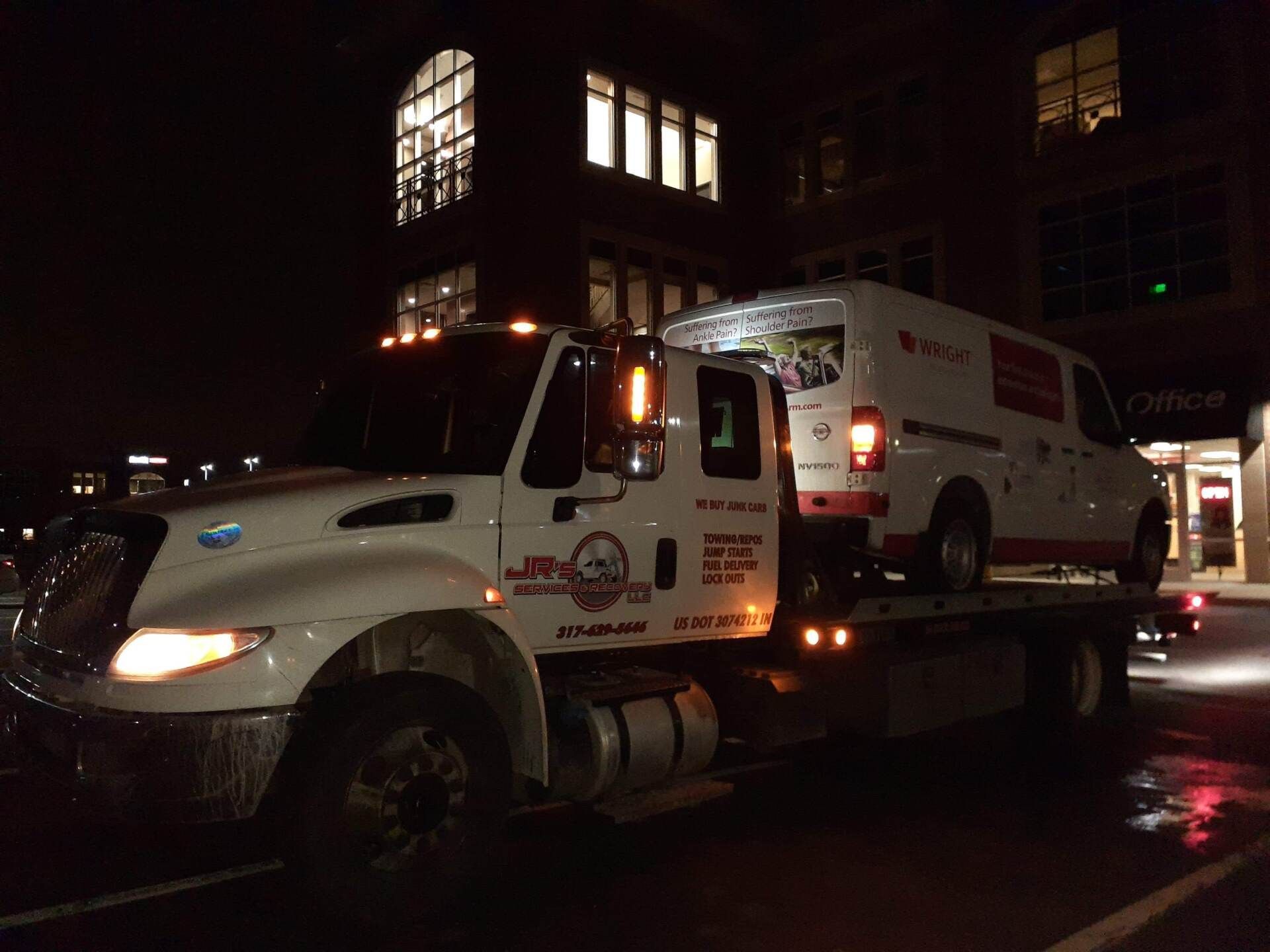 A white tow truck transporting a white delivery van at night near a building with lit windows.