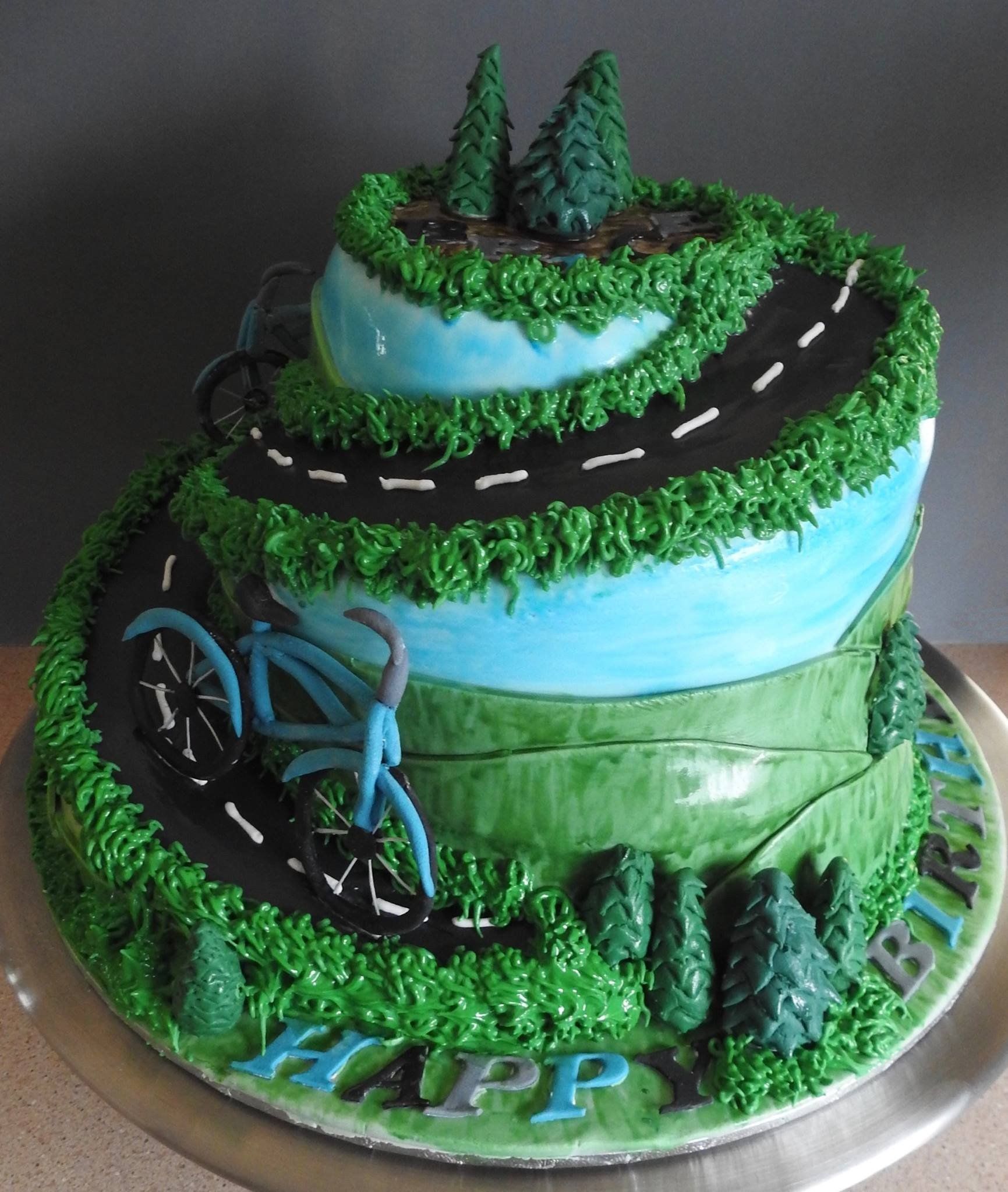 Tierd and shaped birthday cake with fondant hills, trees and two bicycles coming down a road.