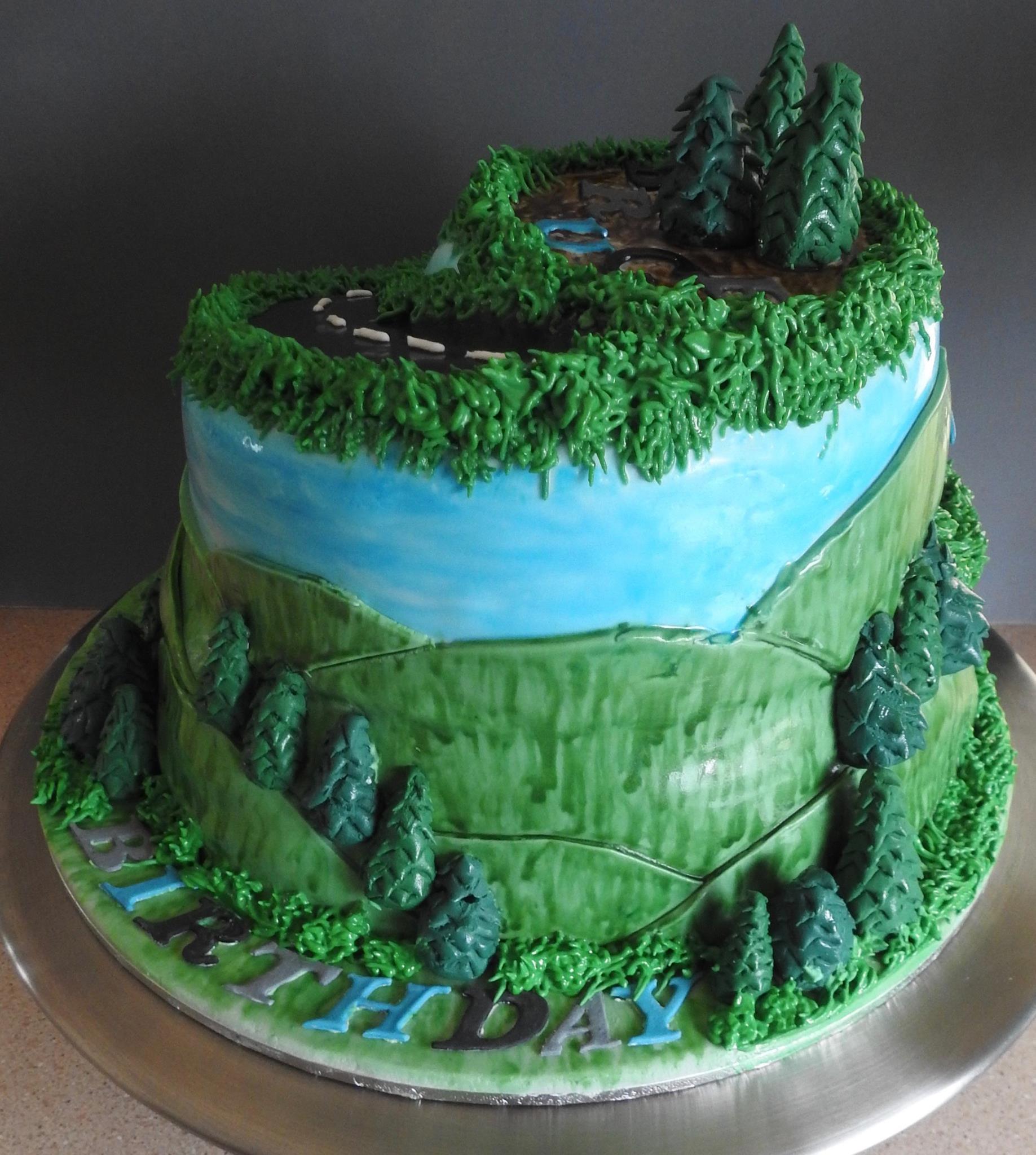 Tierd and shaped birthday cake with fondant hills, trees and two bicycles coming down a road.