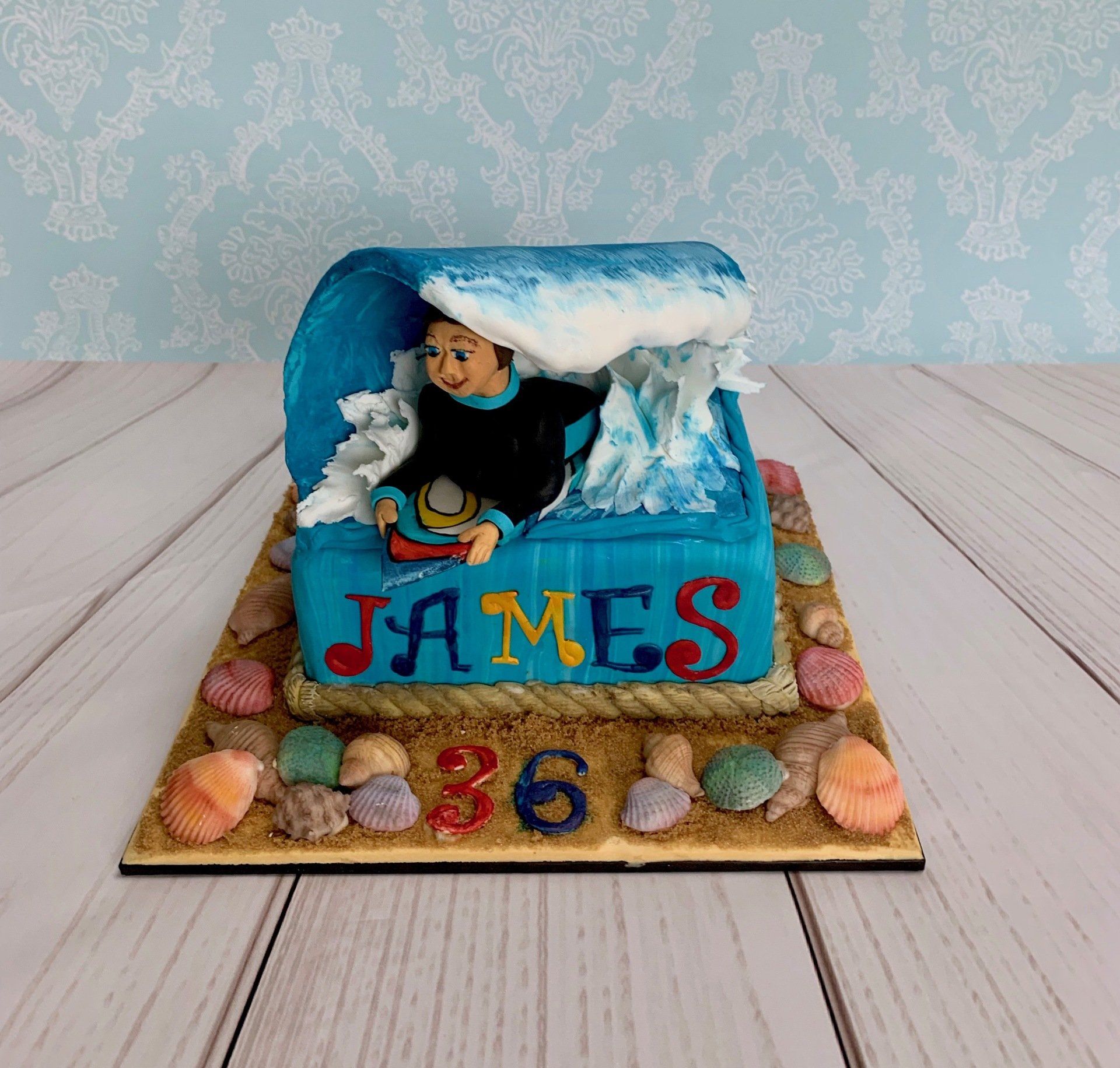 Surfer Themed Cake with sugar sand and shells around the base and handsculpted surfer coming through a wave on top