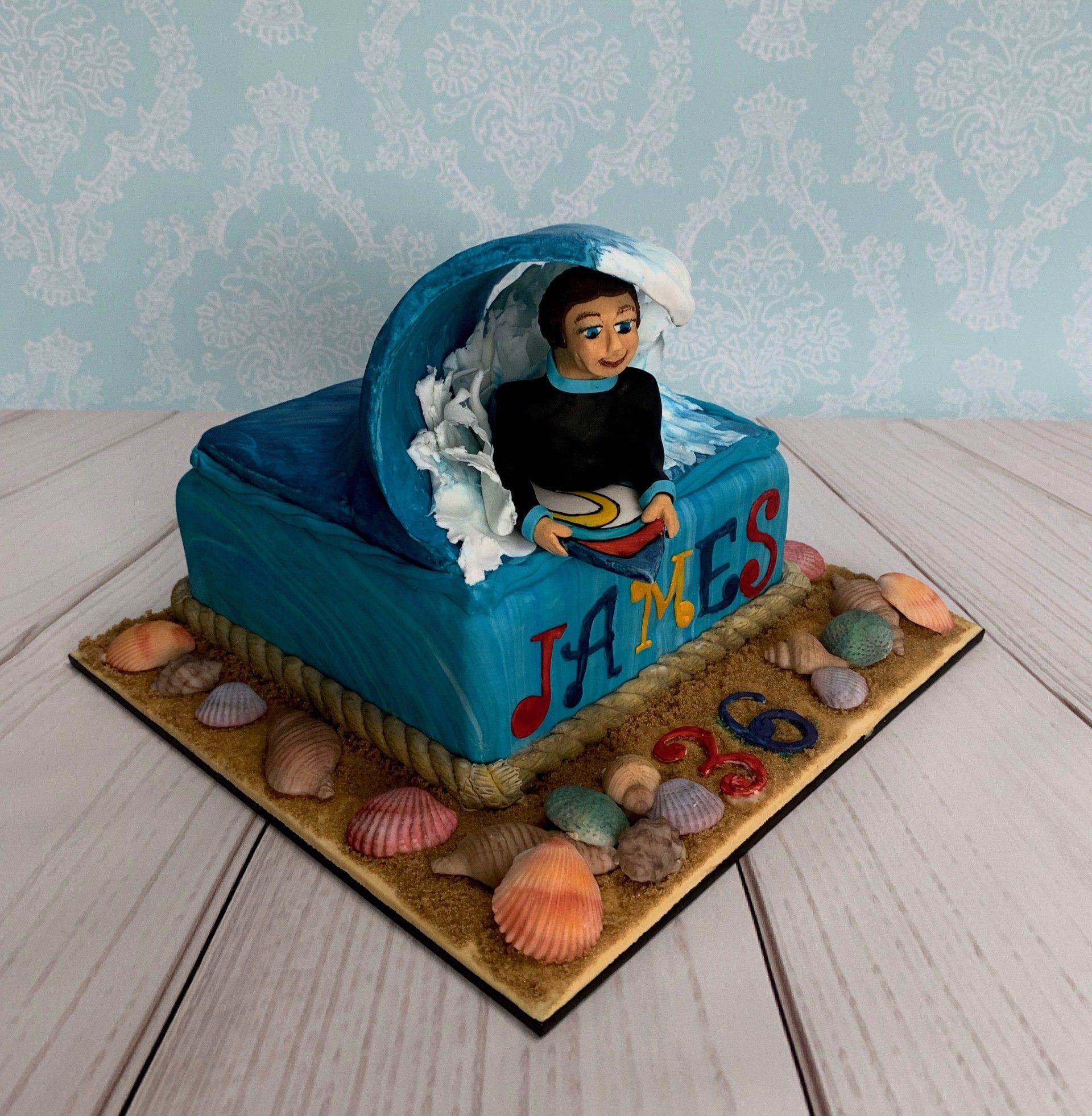 Surfer Themed Cake with sugar sand and shells around the base and handsculpted surfer coming through a wave on top