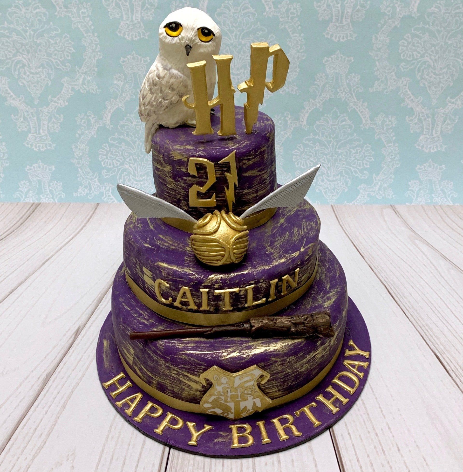3 tier 21st birthday cake in purple and gold with hand sculpted owl, wand, crest, and other magical elements.