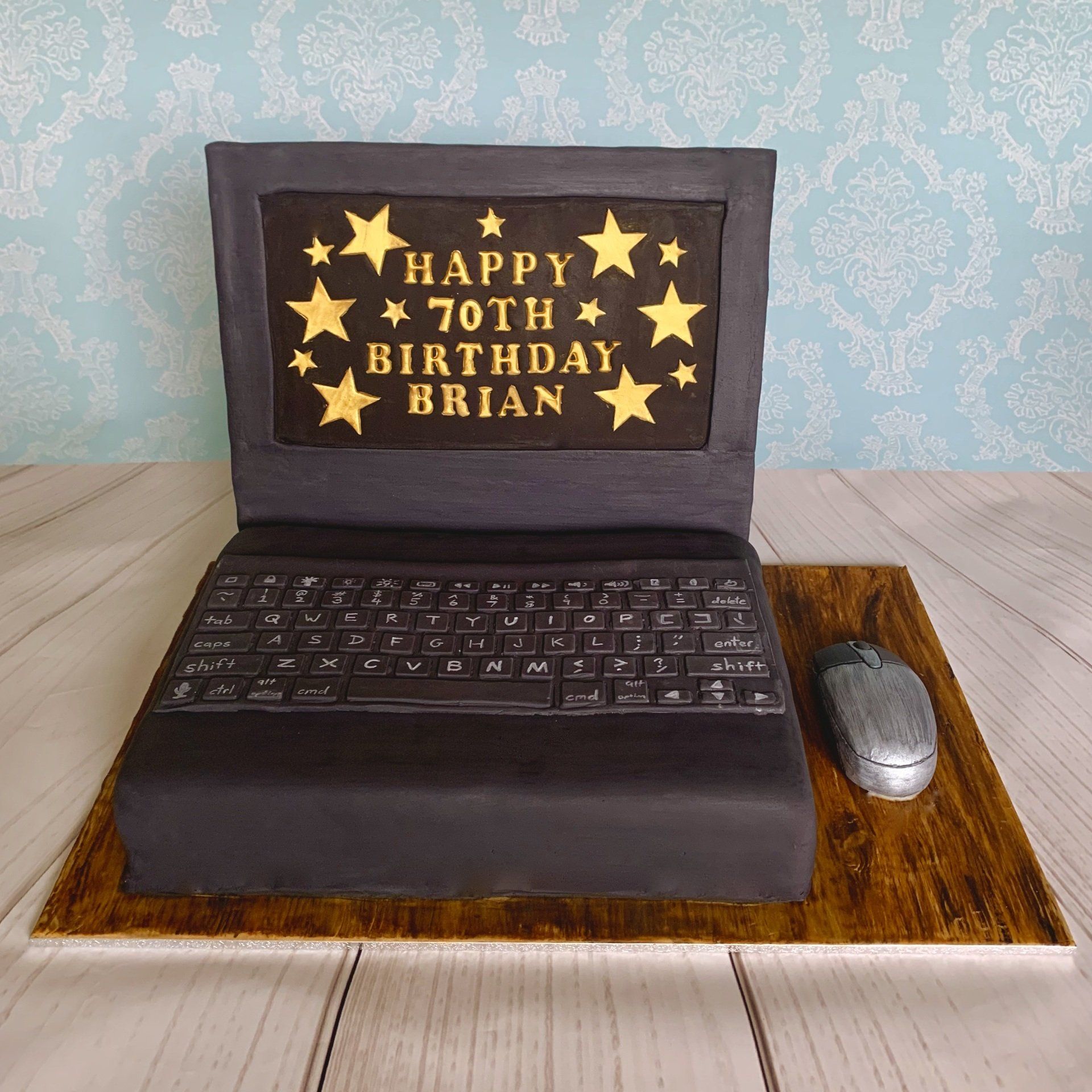 Charcoal grey and black, Computer Laptop shaped cake. With gold writing and stars on the screen, a hand sculpted computer mouse and qwerty keyboard. Cake board covered with a woodgrain fondant effect to look like a desk.