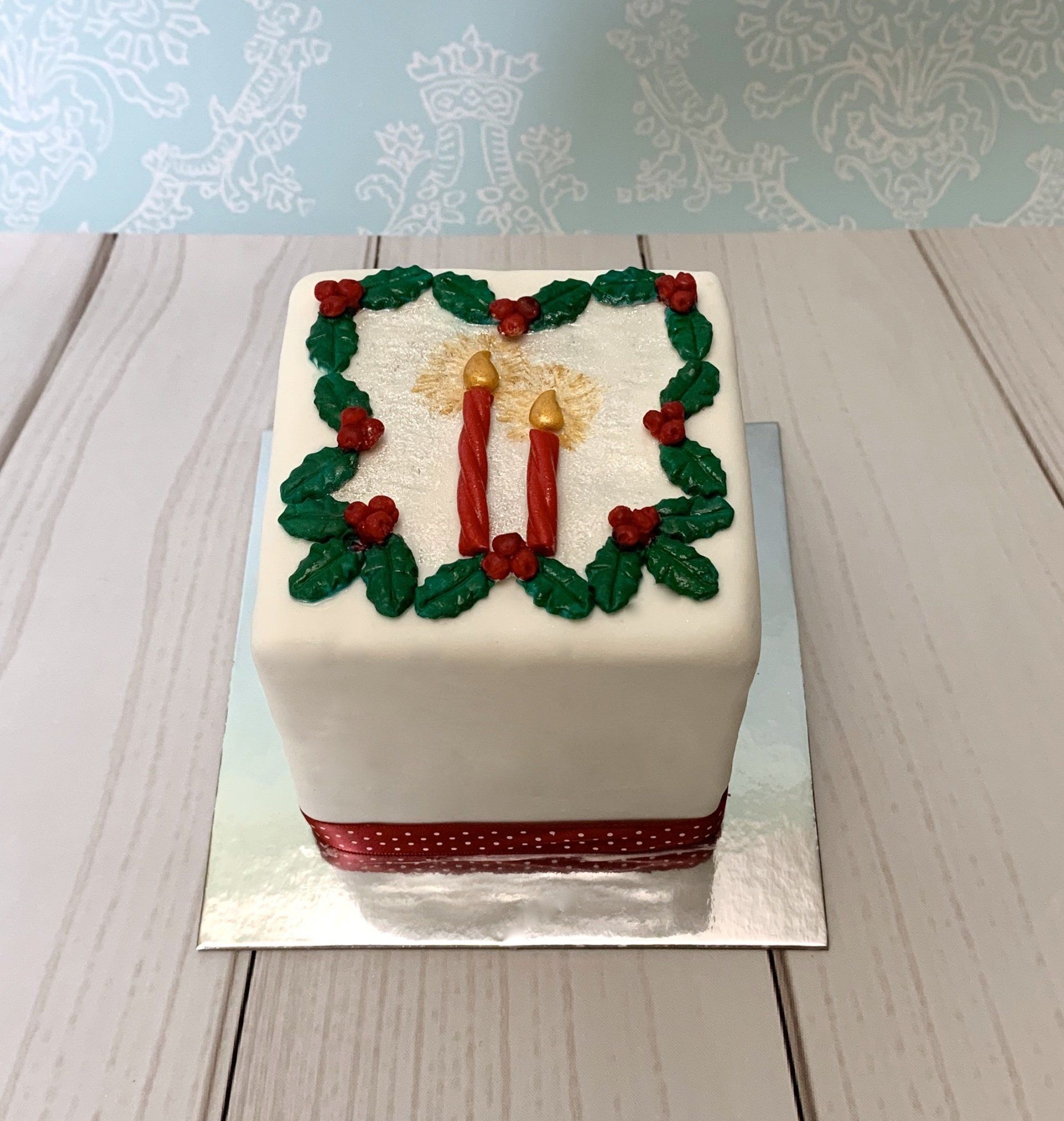 Square Christmas cake with white fondant, red hand crafted sugar candles and sugar holly leaves and berries.