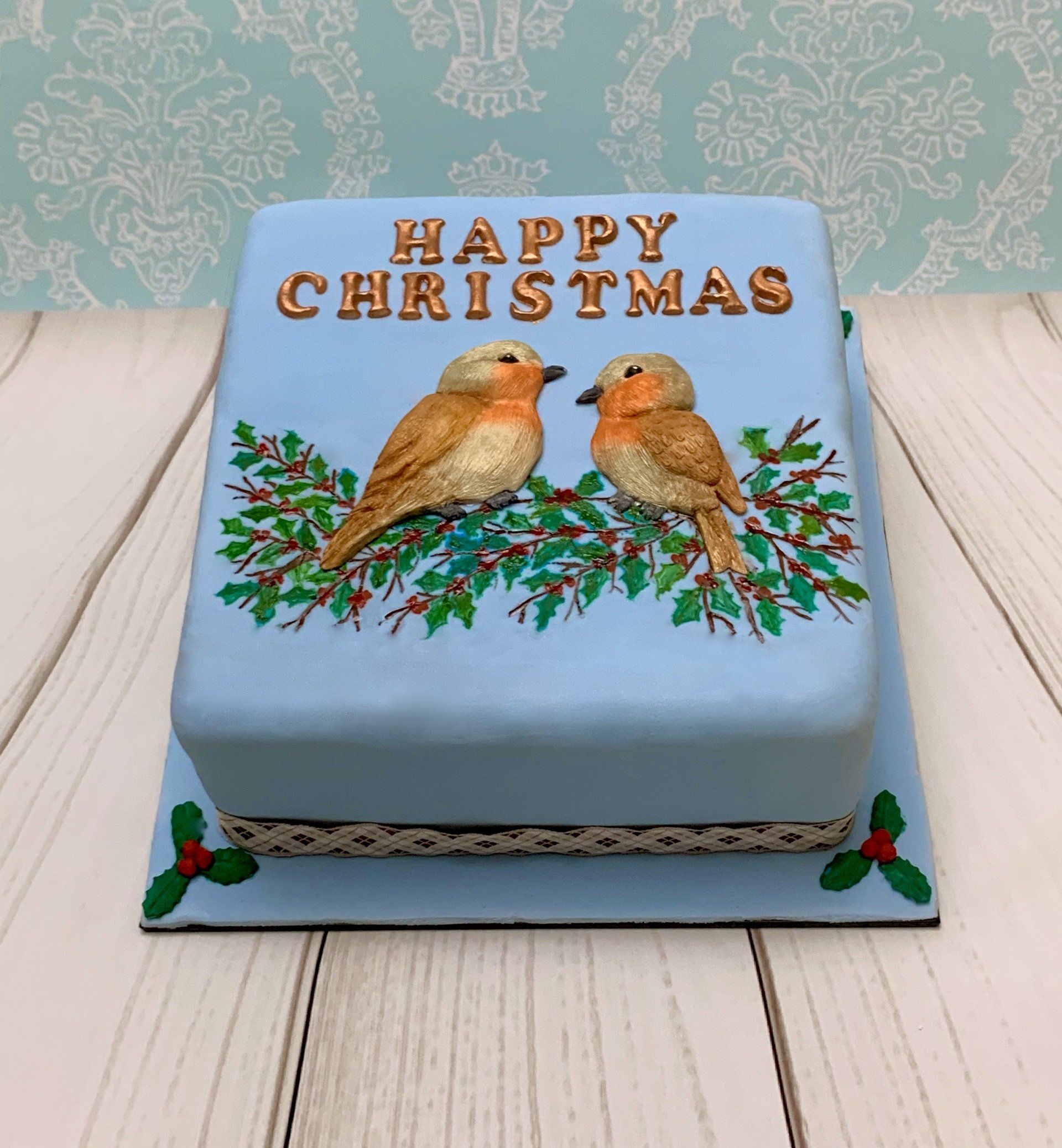 Square Christmas cake with soft blue fondant, rose gold Happy Christmas writing, two gorgeous moulded robins and hand painted holly.