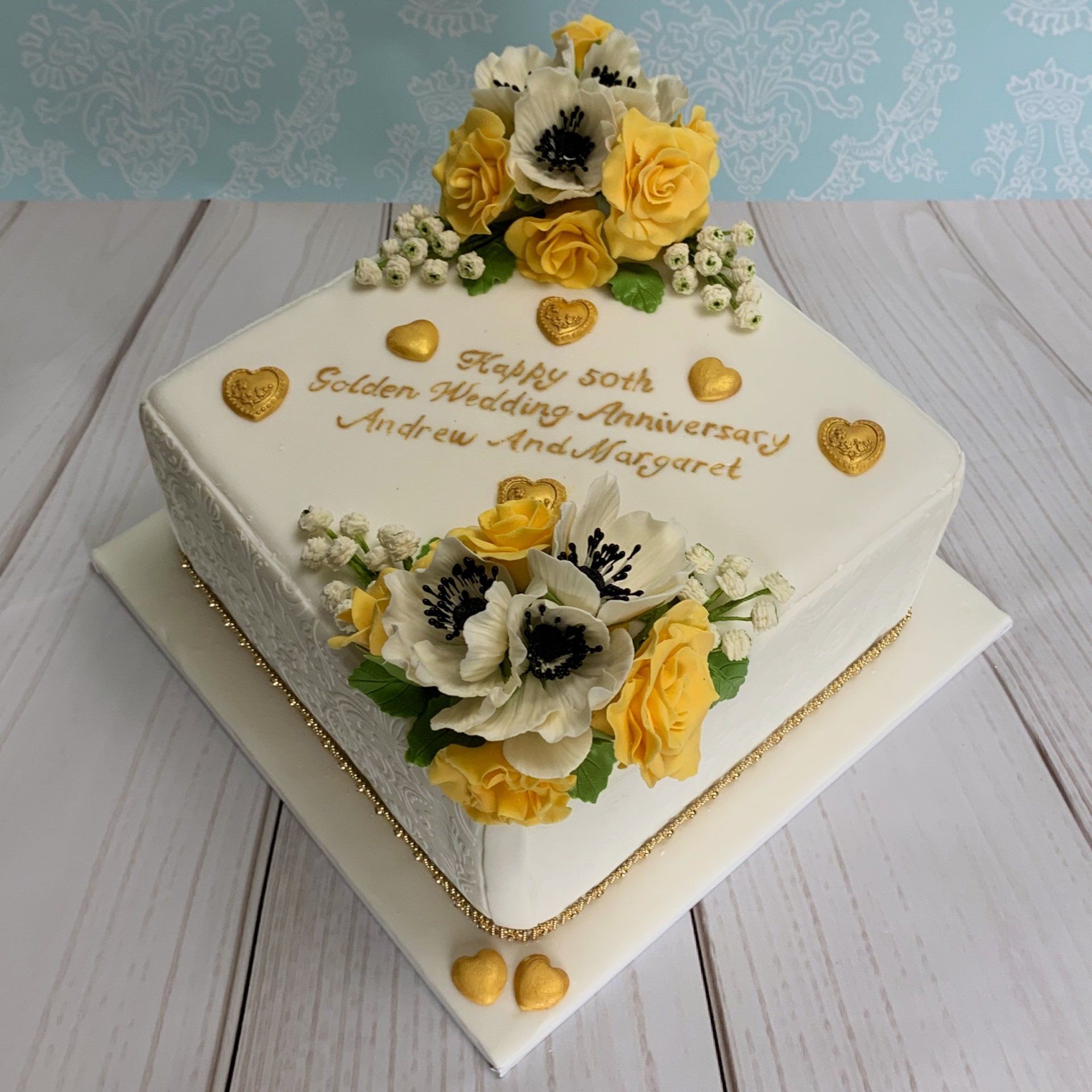 50th golden wedding anniversary cake. Yellow Sugar Roses, White Sugar Anemone Flowers and Sugar Babysbreath arranged on two corners. Gold hand painted writing, Gold hearts and Embossed sides.