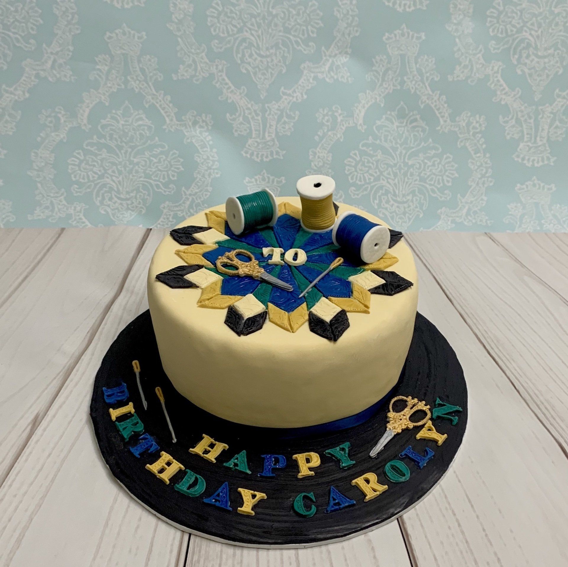 Sewing, patchwork quilt themed cake, with handmade sugar scissors, sewing needles, cotton spools and quilt design.