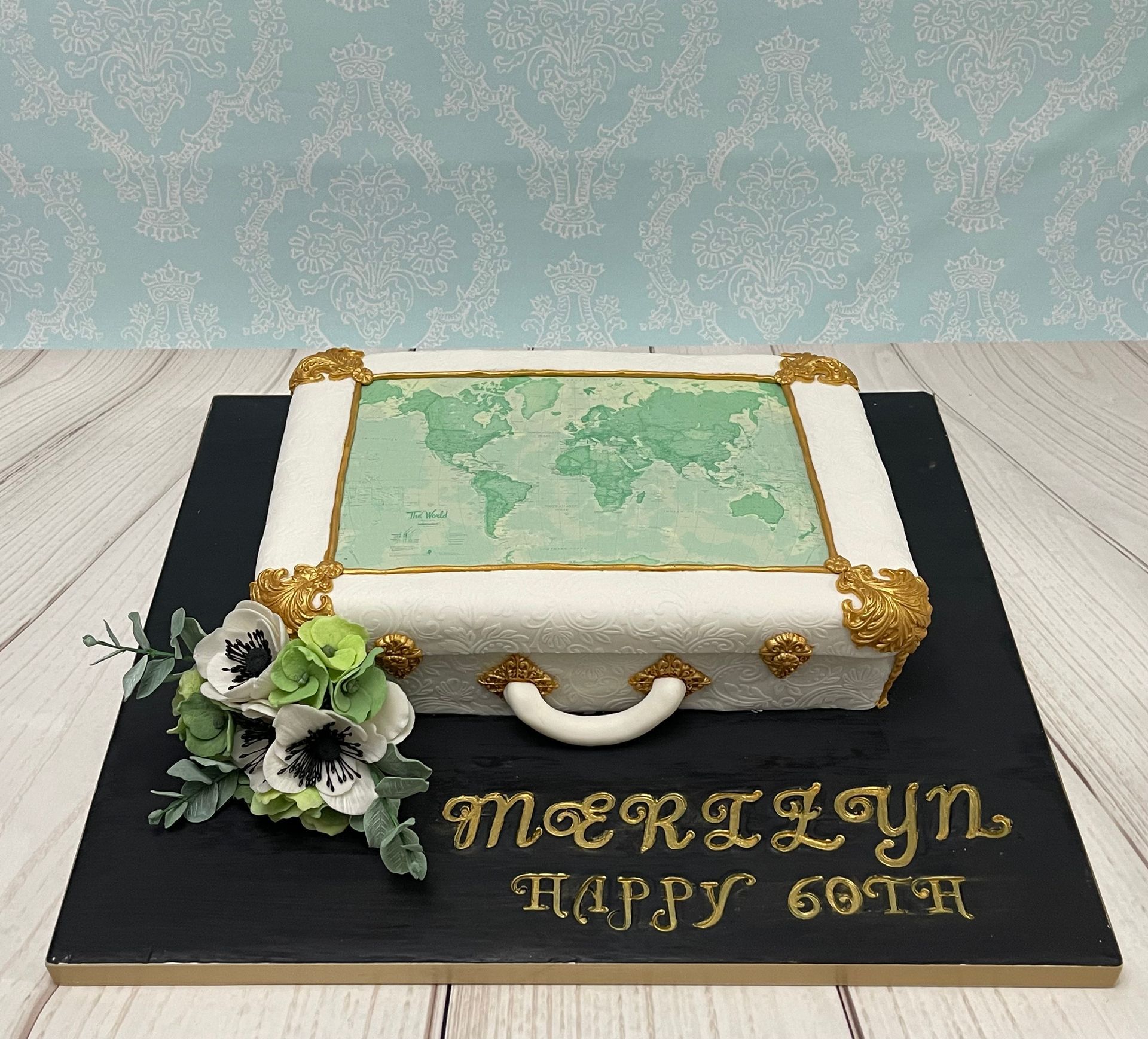 Ornate embossed white suitcase cake with a world map edible print on top, ornate gold fixtures and white and green sugar flowers to decorate. 