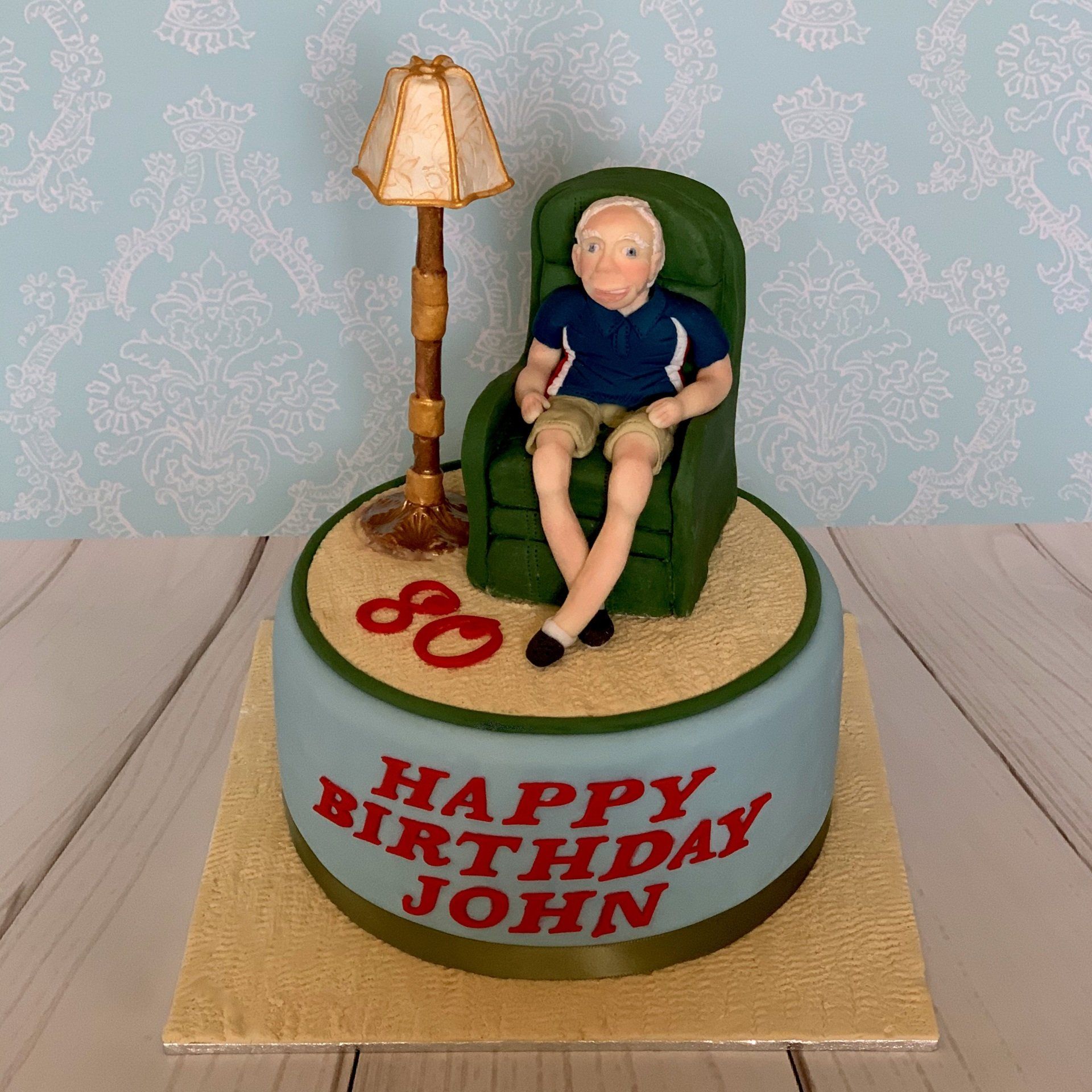 80th Birthday cake with a hand sculpted fondant old man on a green recliner chair and ornate lamp. Along with a carpet textured fondant rug.
