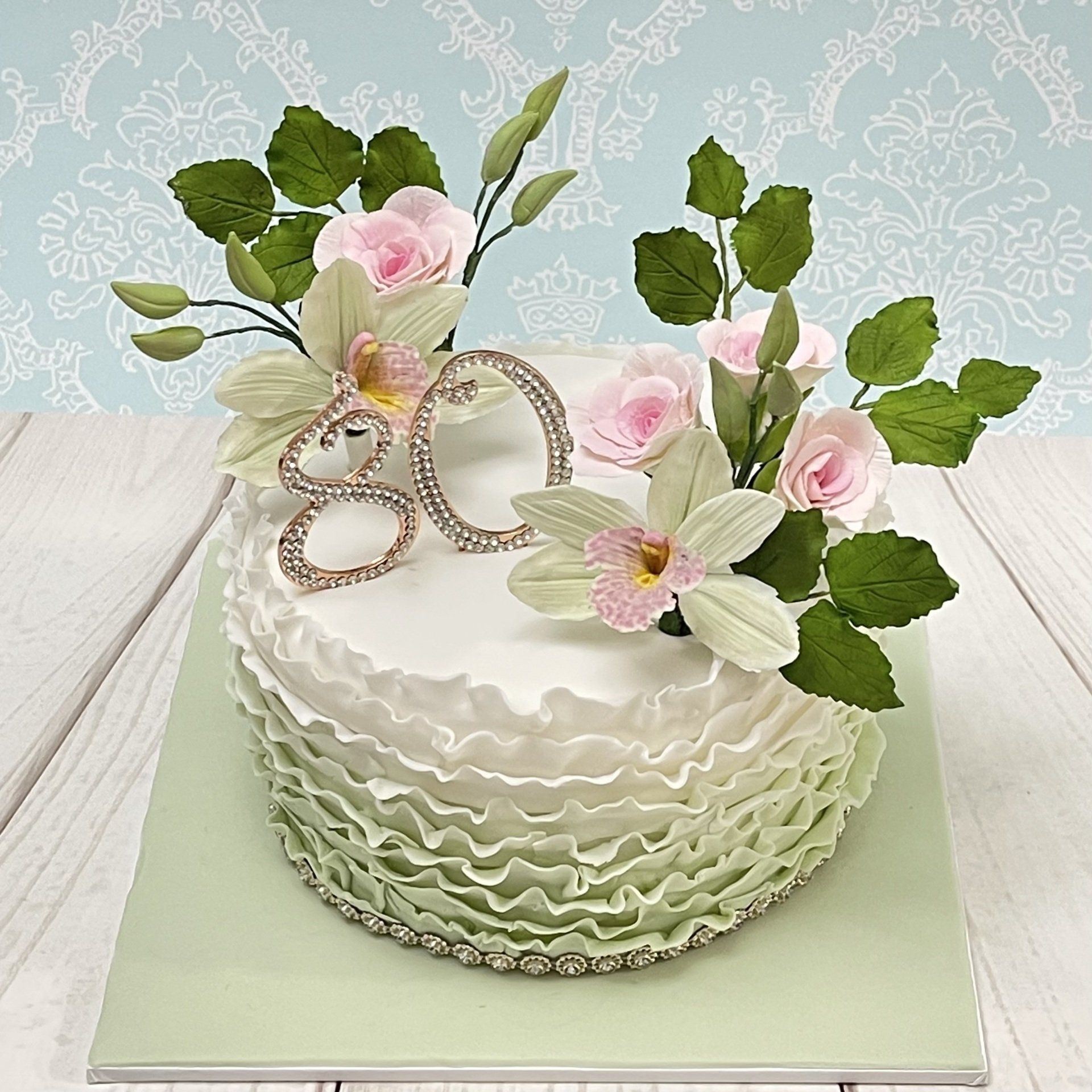 Pretty 80th Birthday Cake with fondant ombre ruffles in white to soft green. Sugar leaves, sugar orchids and sugar roses in soft greens, whites and soft pinks. Plus a rose gold diamante 80 topper.