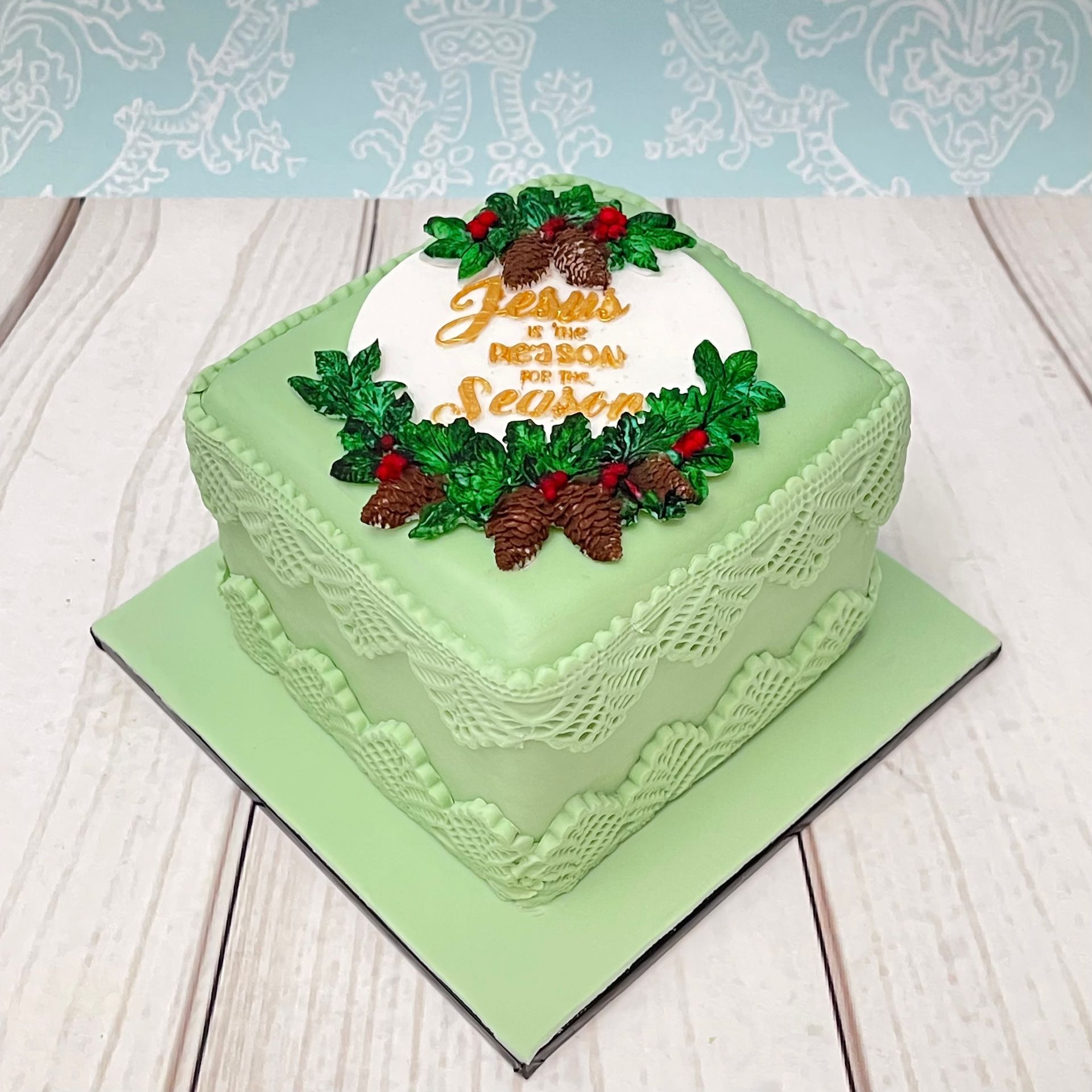 Small Christmas Cake in soft green with soft green fondant lace, Jesus is the reason for the season plaque and Christmas Fondant Foliage. 
