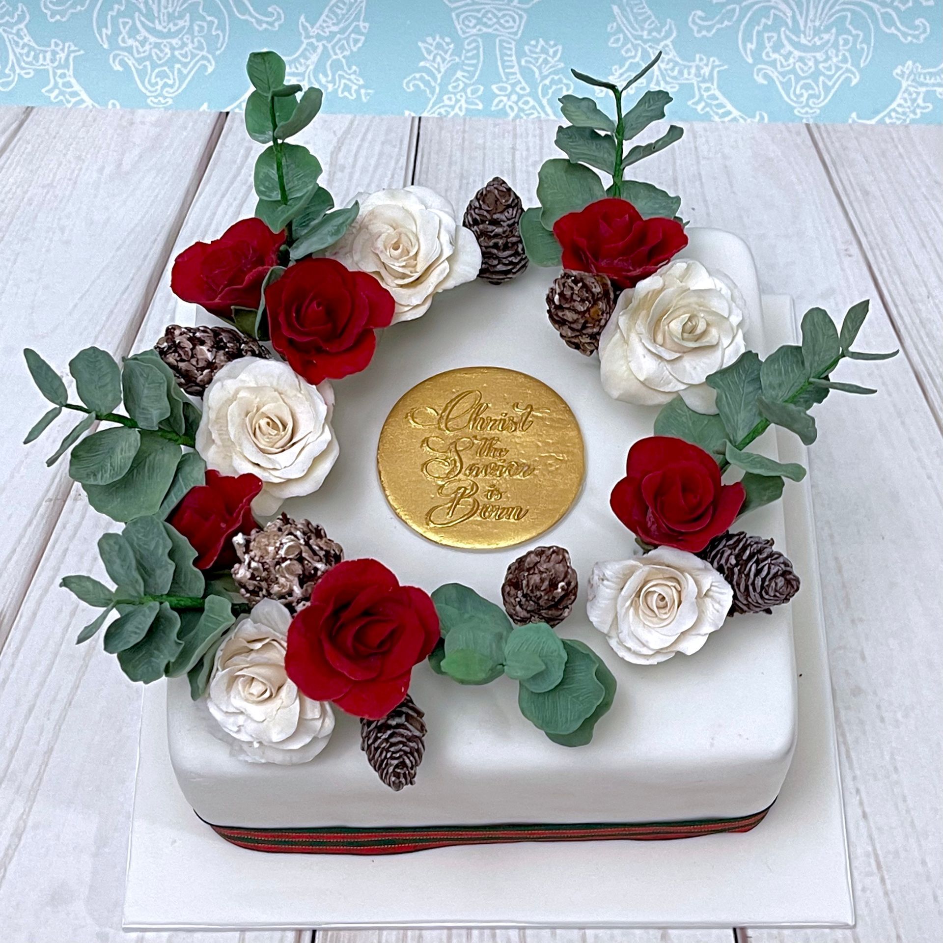 Christmas Wreath Cake With Sugar Roses, Pine Cones and Eucalyptus Leaves with a gold plaque that says 'Christ the Saviour is Born'