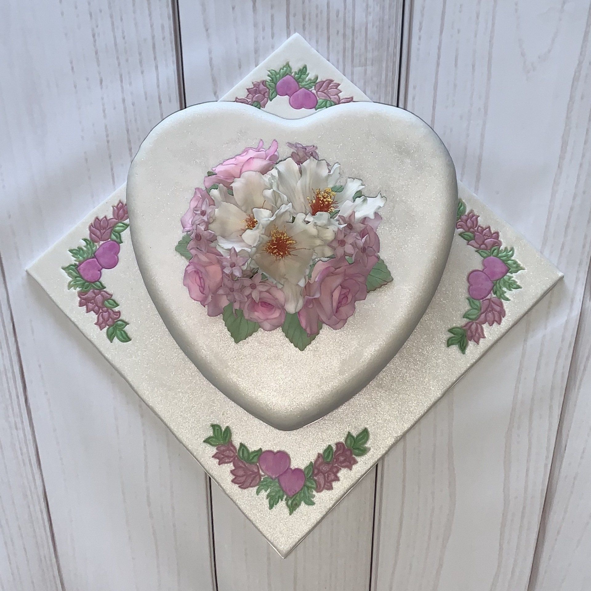 Anniversary heart shaped cake. Pearl fondant, Soft pink and white sugar flowers, pink ribbon around the base and hand painted heart and flower details on the board. Top view.