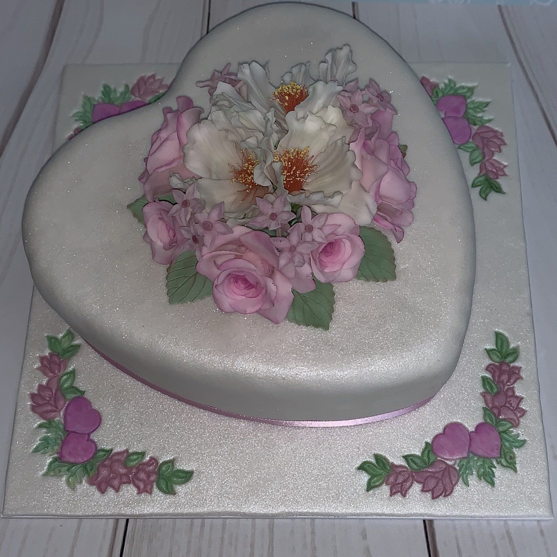 Anniversary heart shaped cake. Pearl fondant, Soft pink and white sugar flowers, pink ribbon around the base and hand painted heart and flower details on the board. Side view.