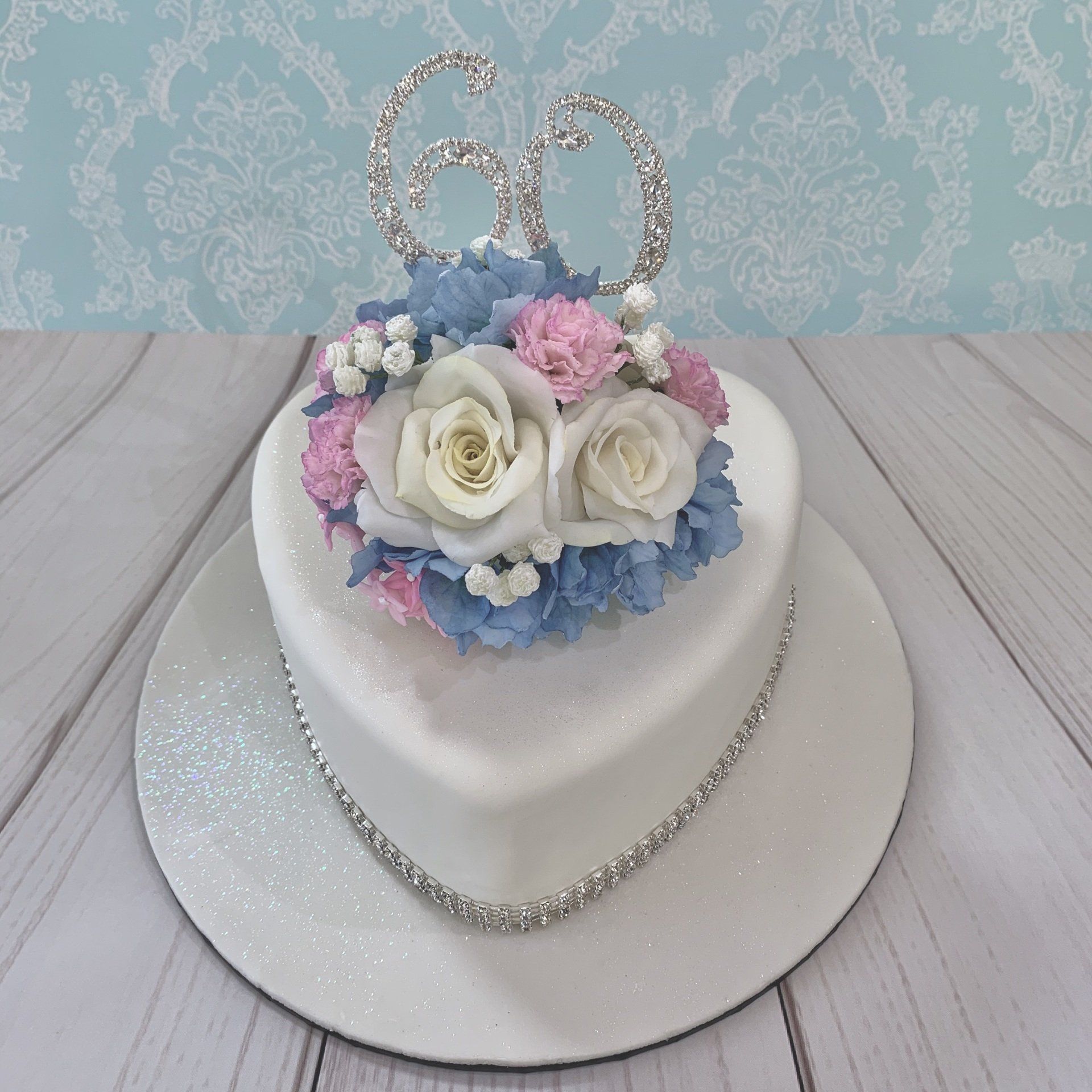 60th Diamond Anniversary heart shaped cake. White shimmer fondant. White sugar roses and sugar baby's breath, , Soft blue sugar Hydrangeas and soft pink sugar carnations. Diamanté 60 and Diamanté trim around the base.