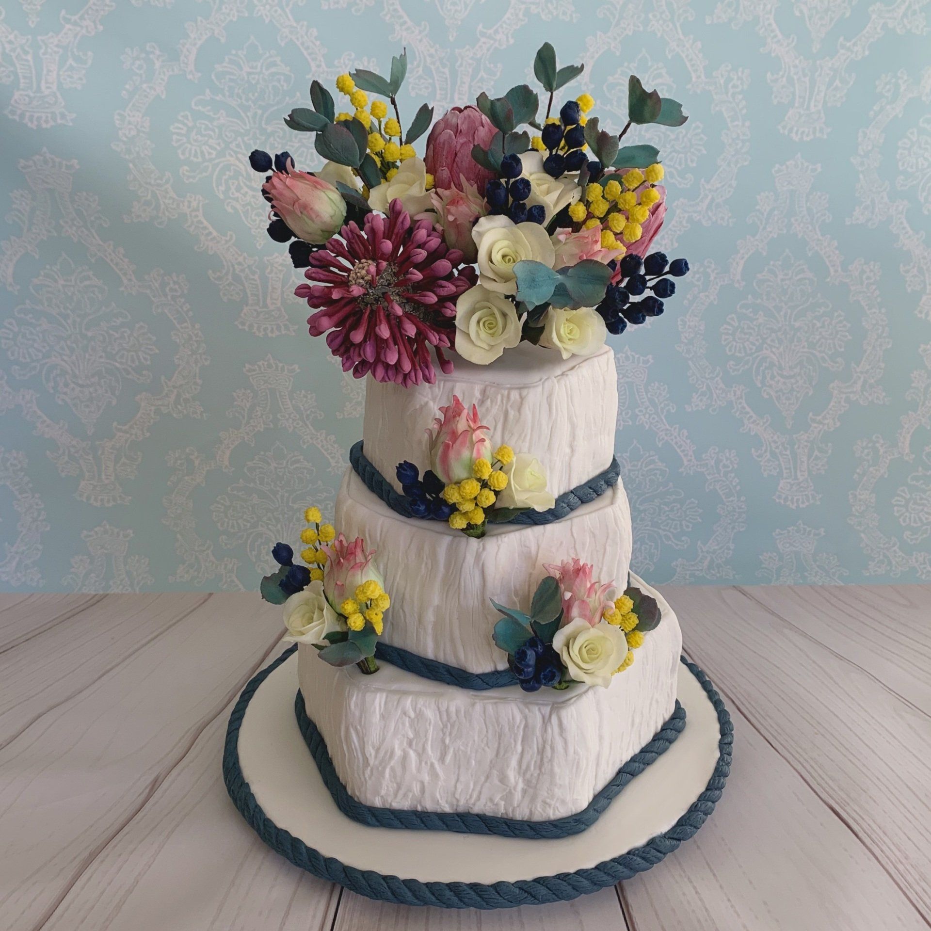 Three tier hexagonal offset wedding cake. White fondant with bark texture, soft navy fondant rope trim around the base and Sugar flower arrangements consisting of Australian native flowers, white roses and blue berries that were all hand sculpted.