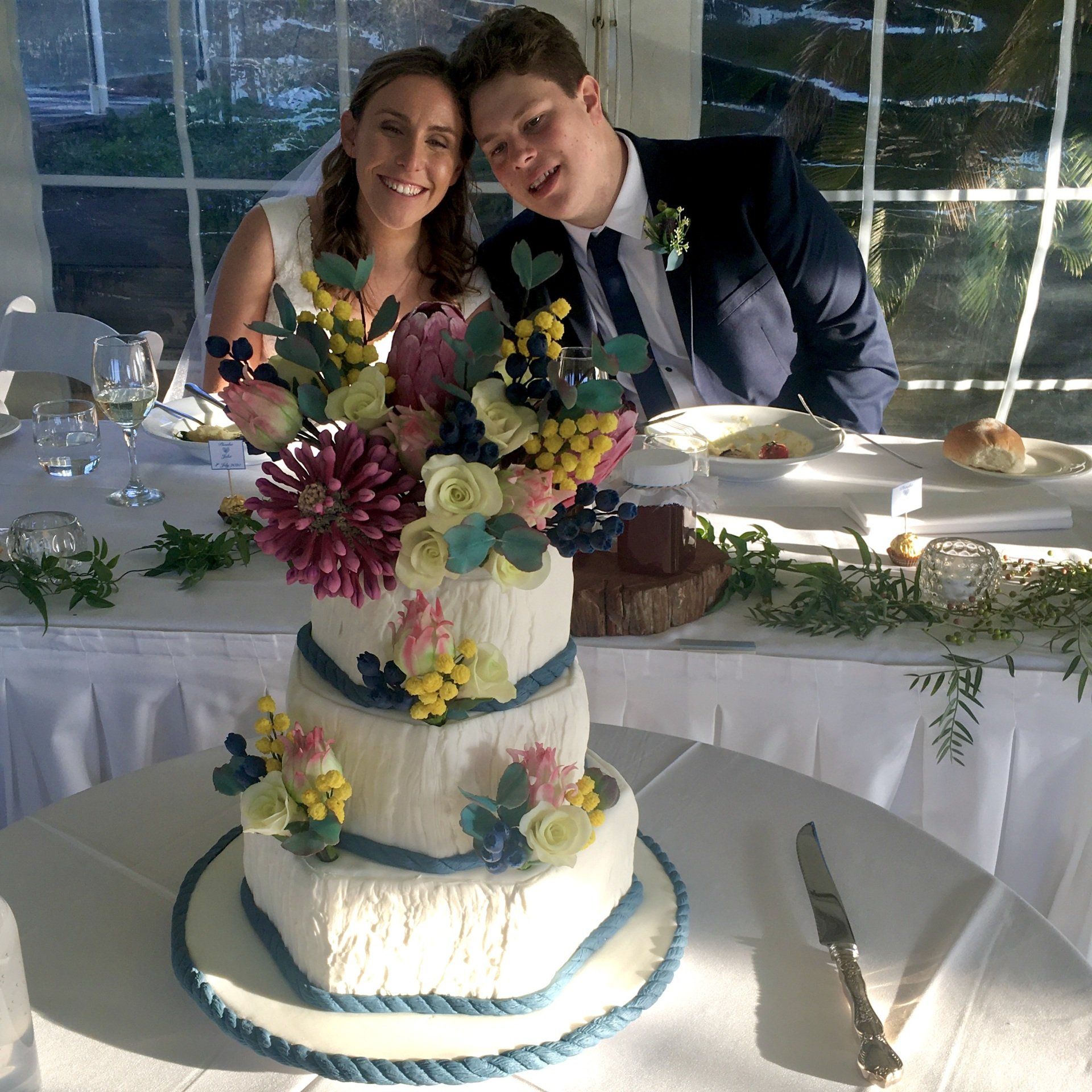 Three tier hexagonal offset wedding cake. White fondant with bark texture, soft navy fondant rope trim around the base and Sugar flower arrangements consisting of Australian native flowers, white roses and blue berries that were all hand sculpted.