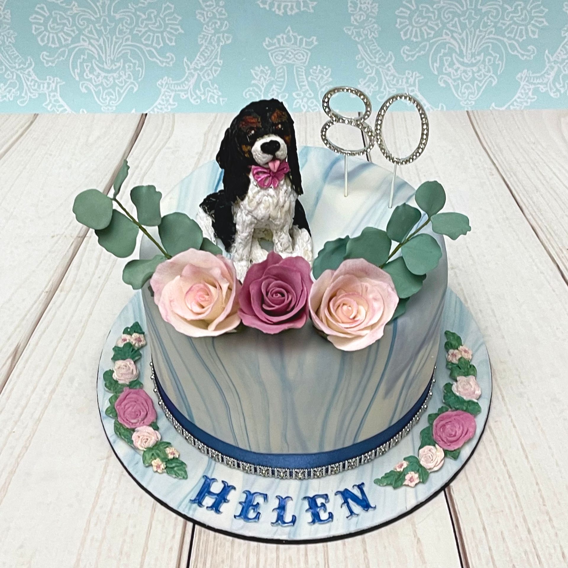 Elegant Birthday Cake with white and blue marbled fondant, blue ribbon and diamanté trim, diamanté 80, a cute hand sculpted fondant Cavalier King Charles Spaniel with pink bow floral moldings name and handmade sugar roses and sugar eucalyptus leaves 
