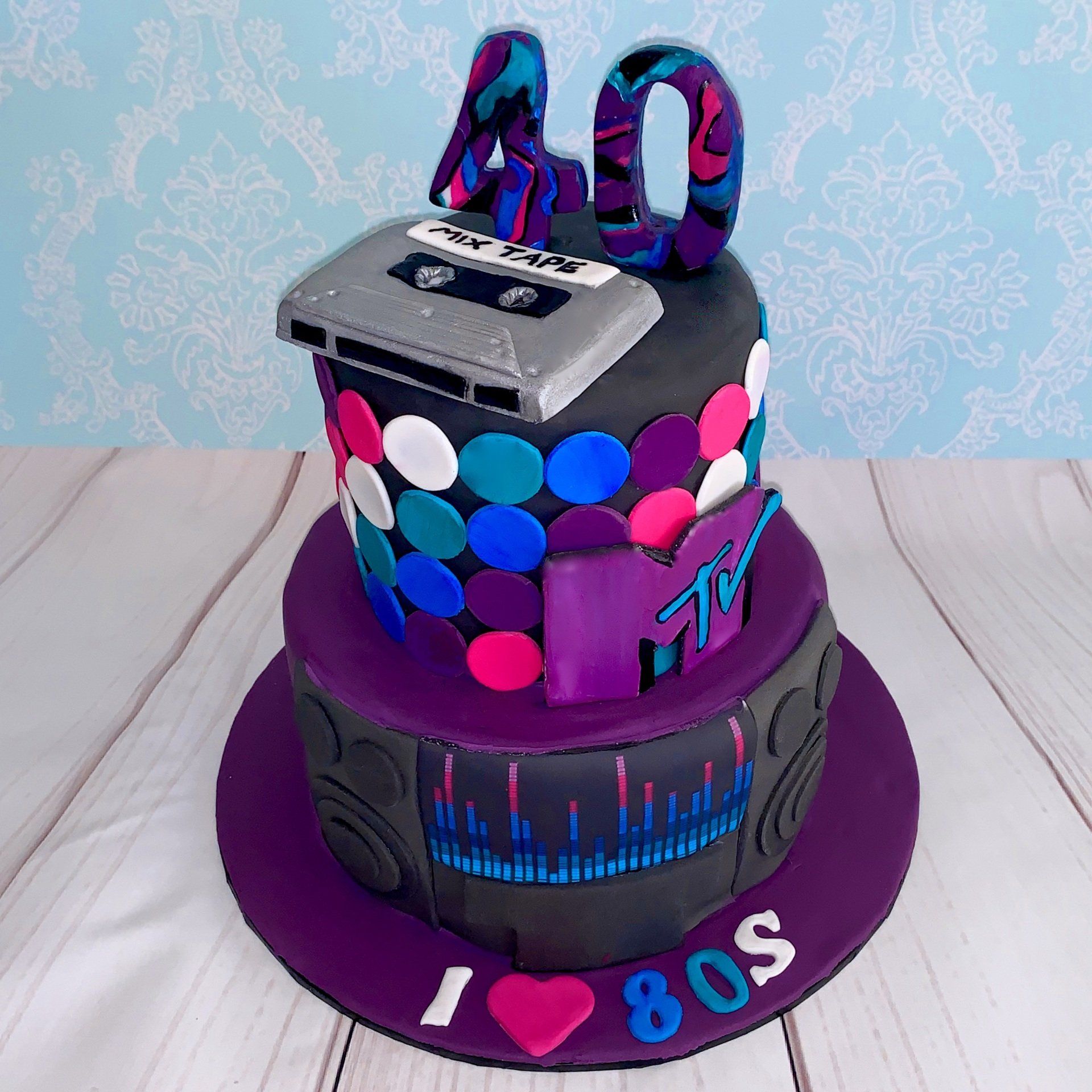80's themed cake with bright purple, pink, blue, teal, black and white  fondant. A fondant cassette mix tape, and other 80's logos.
