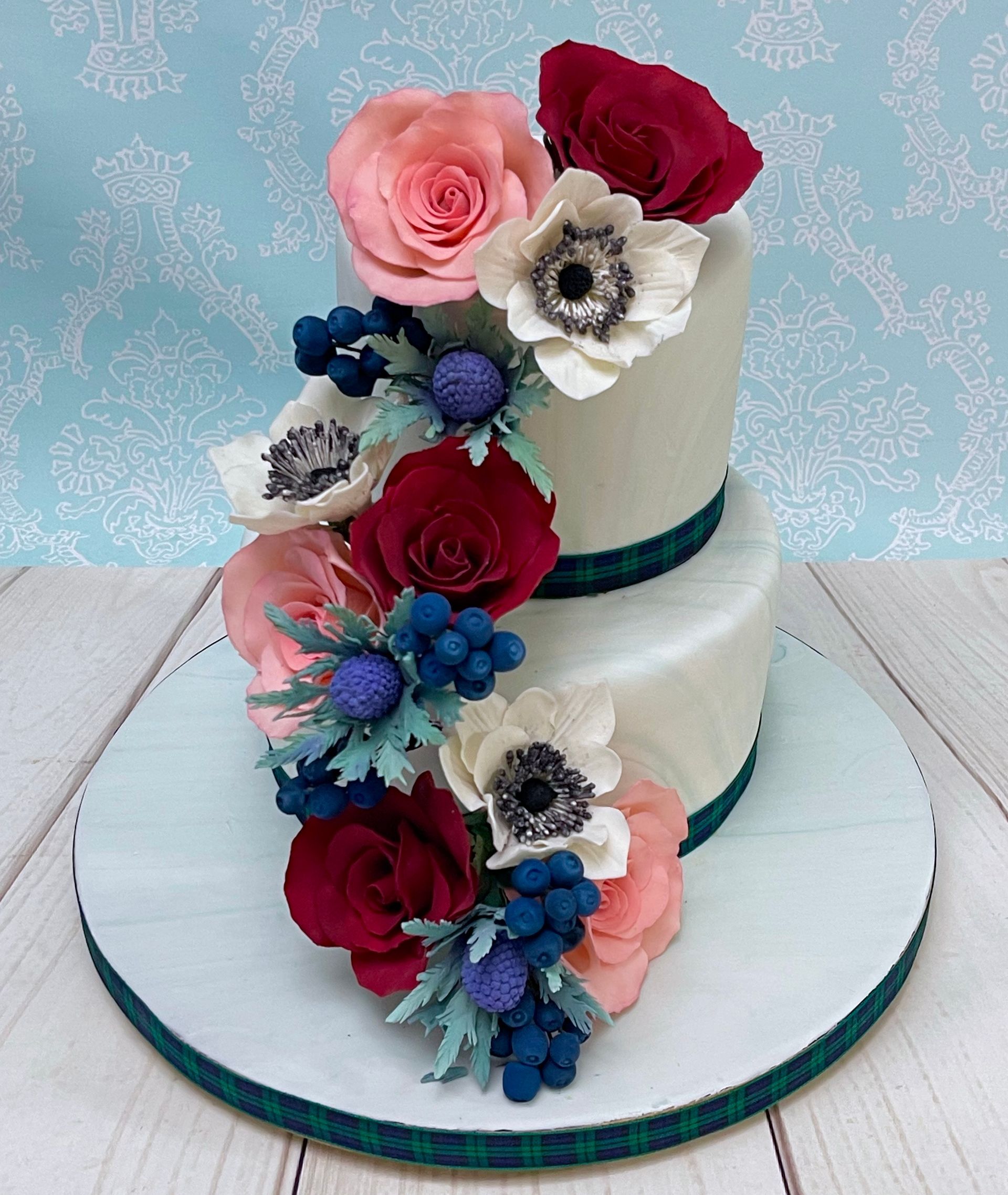 Thank you and Farewell cake Port Macquarie. Two tier with soft marbled fondant, Tartan ribbon trim. Handmade Cascading Sugar roses in red and pinkish apricot, white Sugar Anenomes, Sugar Blue berries and sugar Sea holly thistles. 
