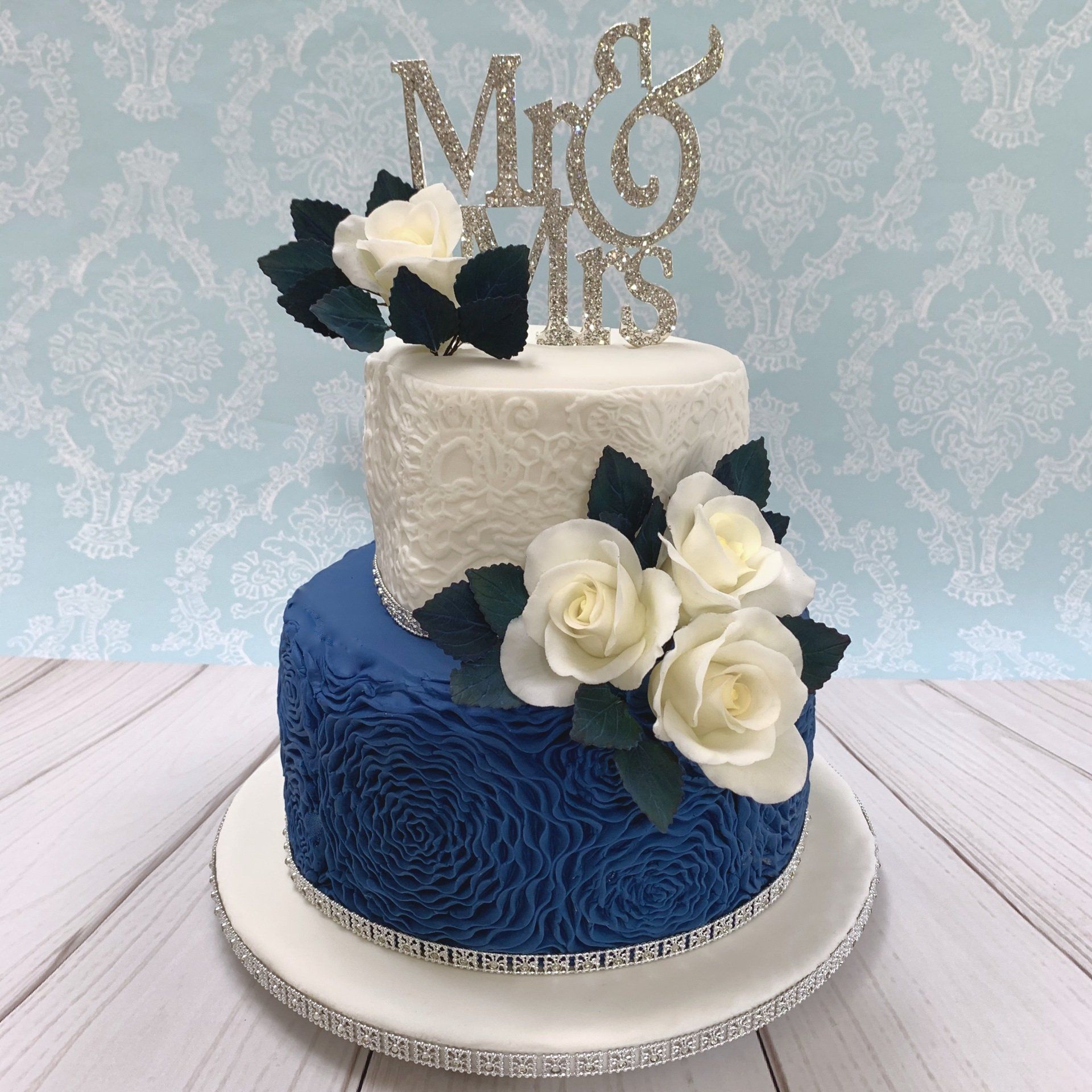 Two tier Wedding cake with teal fondant rosettes around the base, lave impression around the top, white sugar roses with teal sugar leaves and a diamanté Mr & Mrs topper.