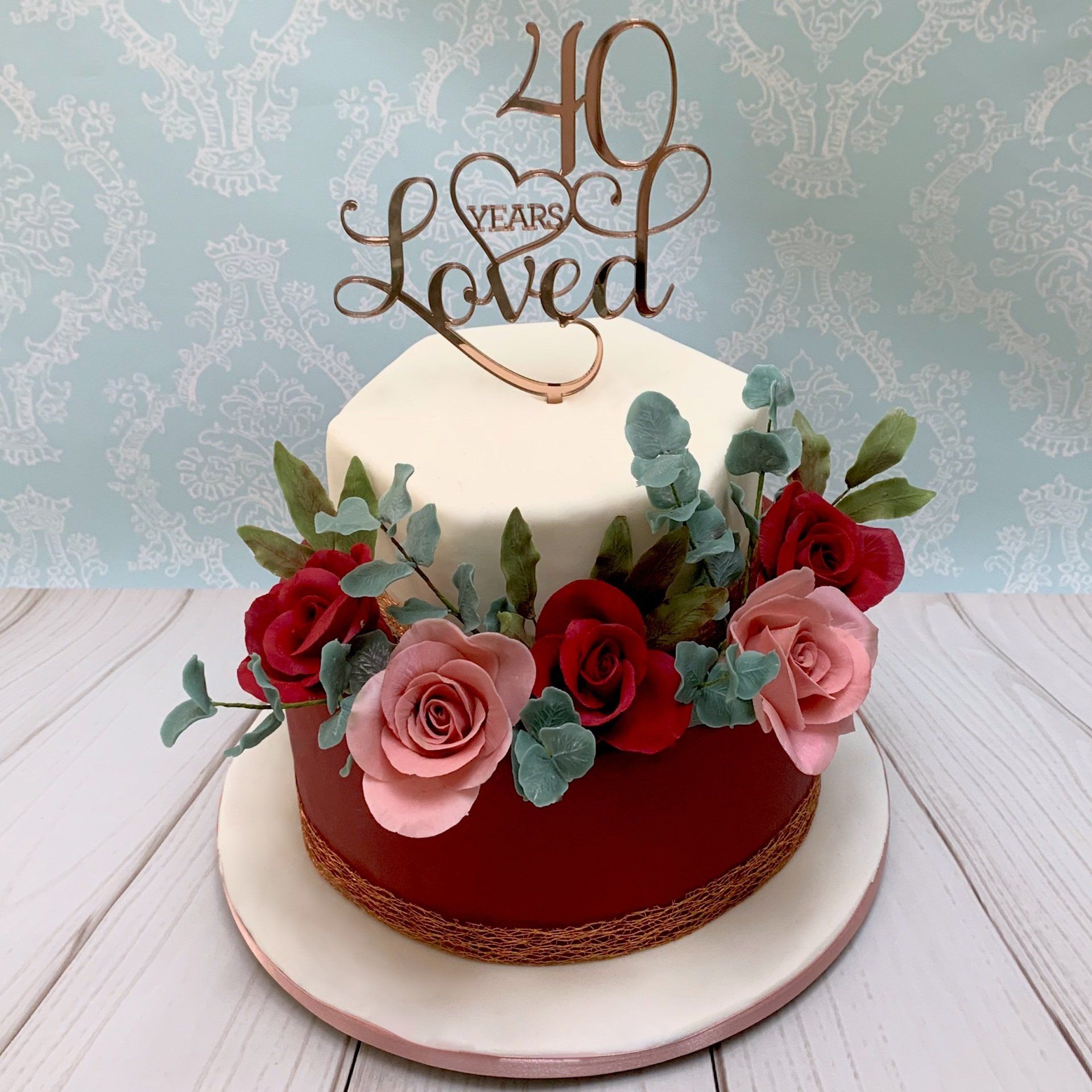 40th Ruby Wedding Anniversary two tiered cake.  White fondant hexagonal top tier, Burgundy fondant round base tier. Rose Gold trim and Anniversay topper. Deep red and pink sugar roses and sugar leaves.